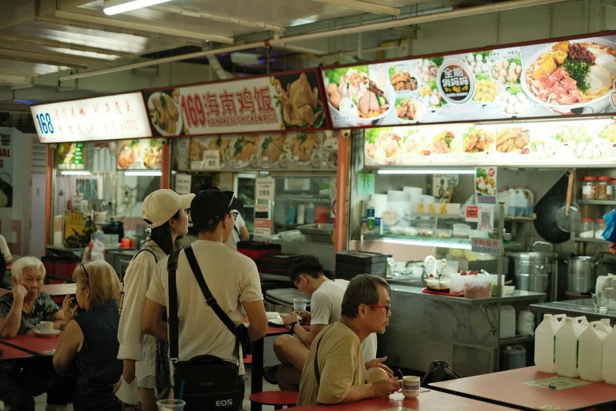 People queuing and dining at a brightly lit Singapore hawker centre with food stalls and menu boards overhead.