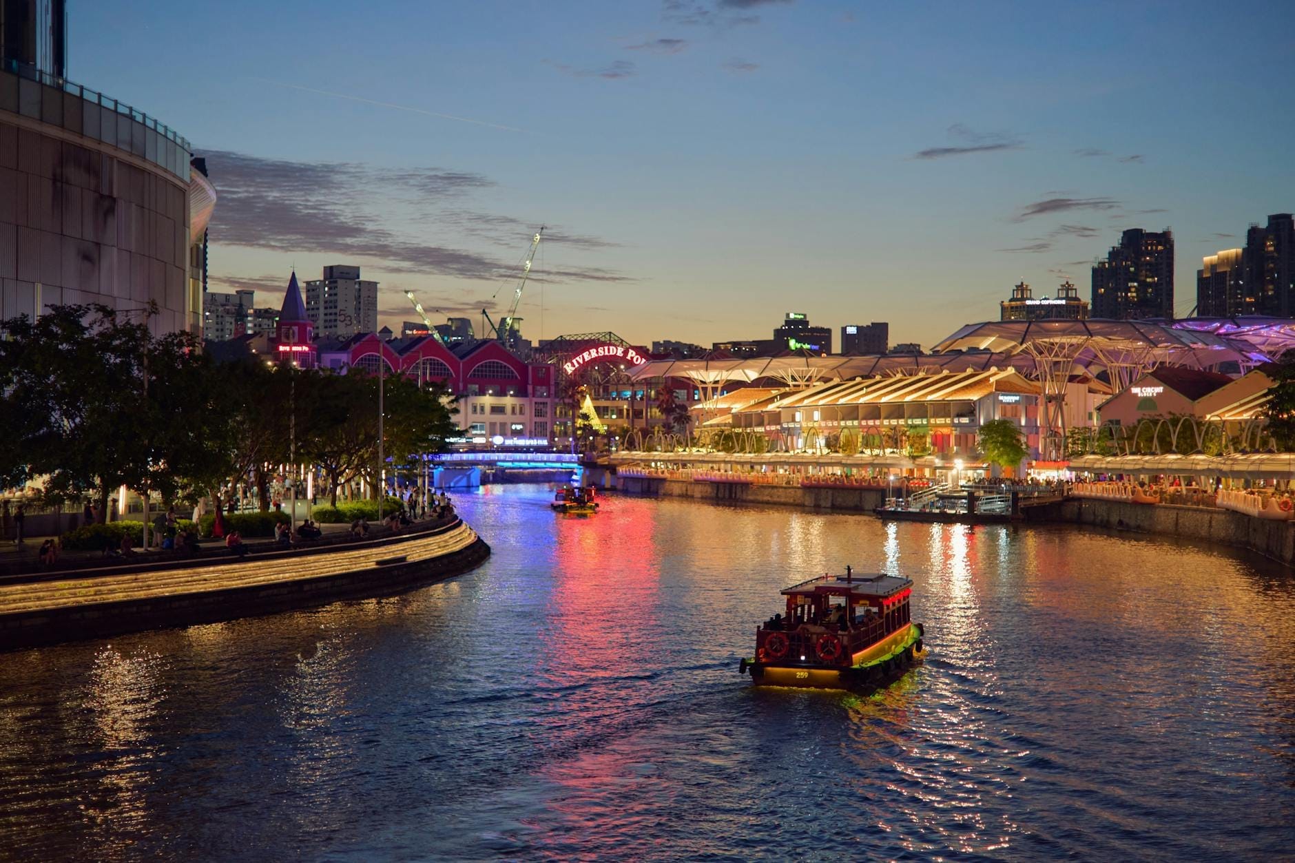 เรือบัมโบ๊ทดั้งเดิมล่องแม่น้ำสิงคโปร์ยามค่ำ ท่ามกลางแสงสีสดใสของ Clarke Quay และสกายไลน์เมืองทันสมัยที่สร้างบรรยากาศริมน้ำคึกคัก