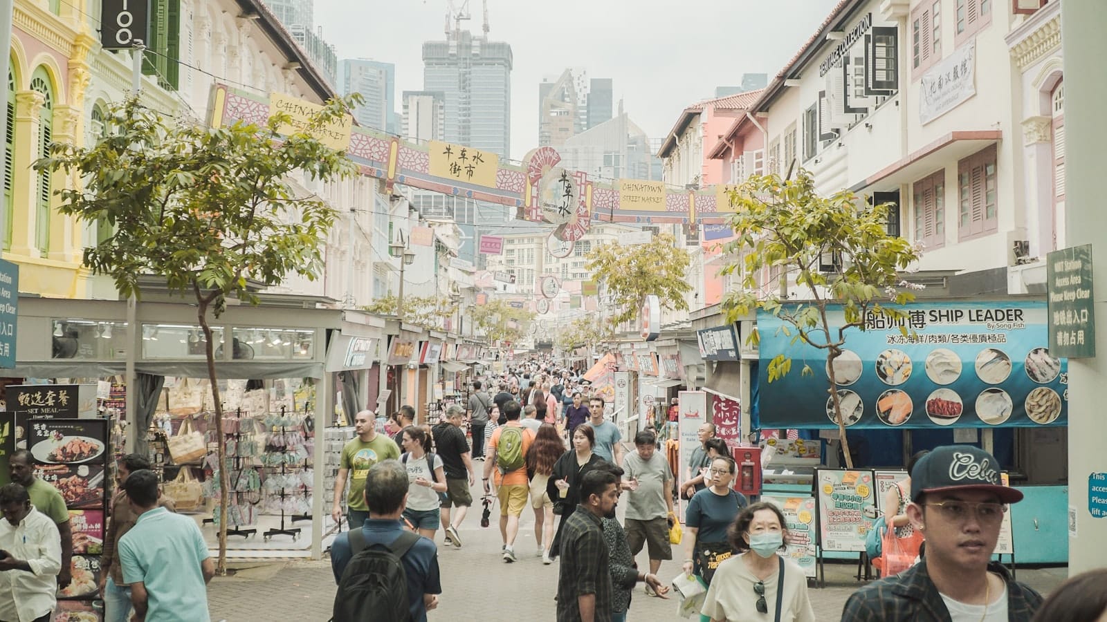Movimentado mercado de rua em Singapura com fachadas coloridas, multidão de compradores e arranha-céus ao fundo num dia ensolarado.