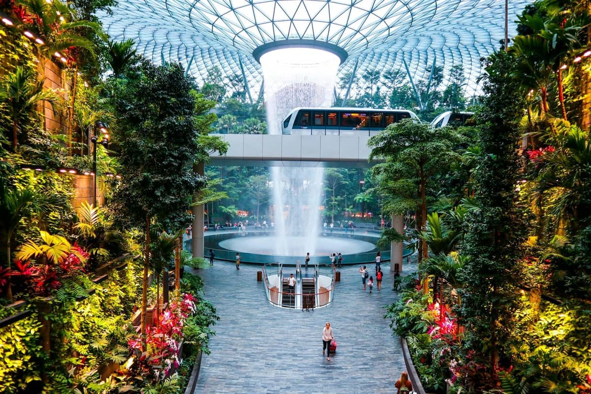 Wide view of Jewel Changi Airport's indoor waterfall surrounded by lush greenery, open walking area, and the connecting skytrain track overhead.