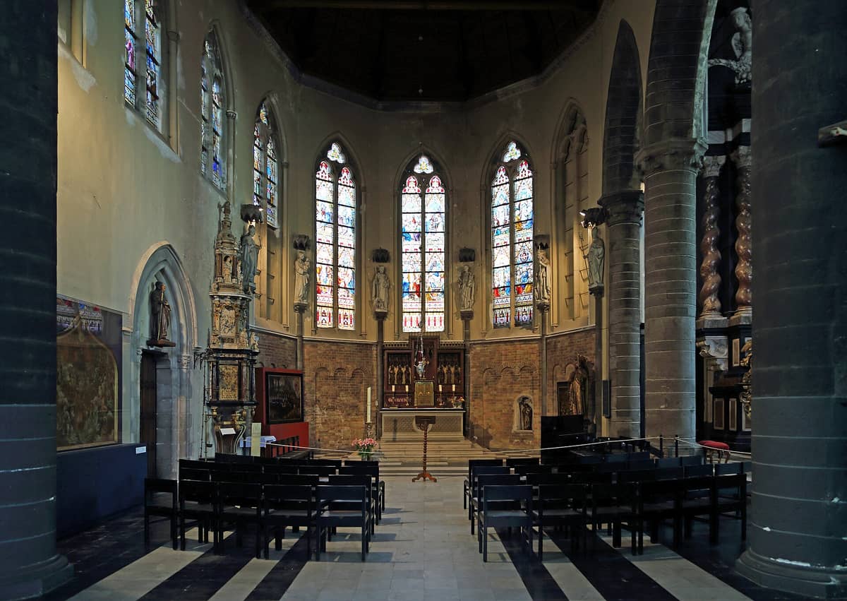Interior of Sint-Jakobskerk in Bruges showing stained glass windows, ornate altarpiece, Flemish artworks, and dark rows of benches in a light-filled chancel.