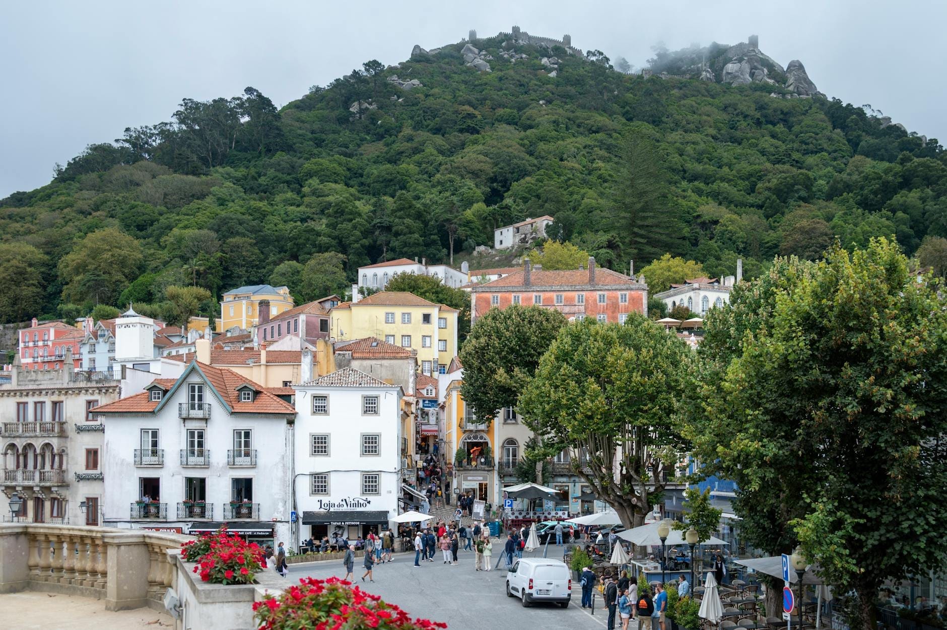 Vue du centre-ville de Sintra avec ses bâtiments colorés, des piétons, des voitures et le Castelo dos Mouros sur la colline boisée.