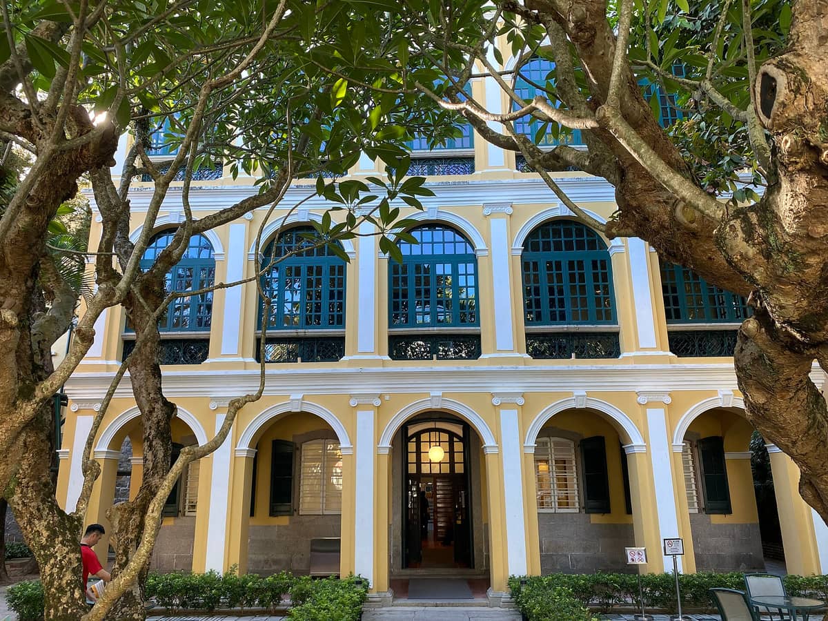 Fassade der Sir Robert Ho Tung Library in Macau mit kolonialen gelben Bögen und grünen Fensterläden, eingerahmt von üppigem Baumgrün im Vordergrund.