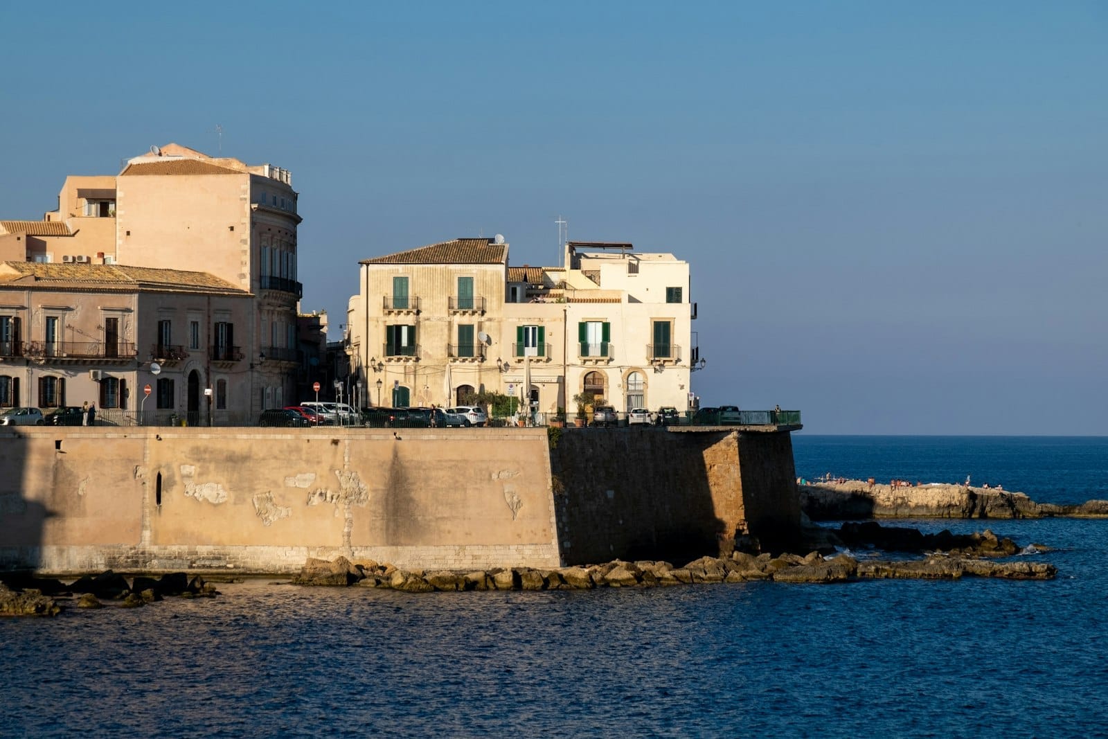 Blick auf die historischen Uferbauten von Ortigia in Siracusa, die sich an einem sonnigen Tag über Steinmauern erheben, mit dem blauen Mittelmeer im Vordergrund.
