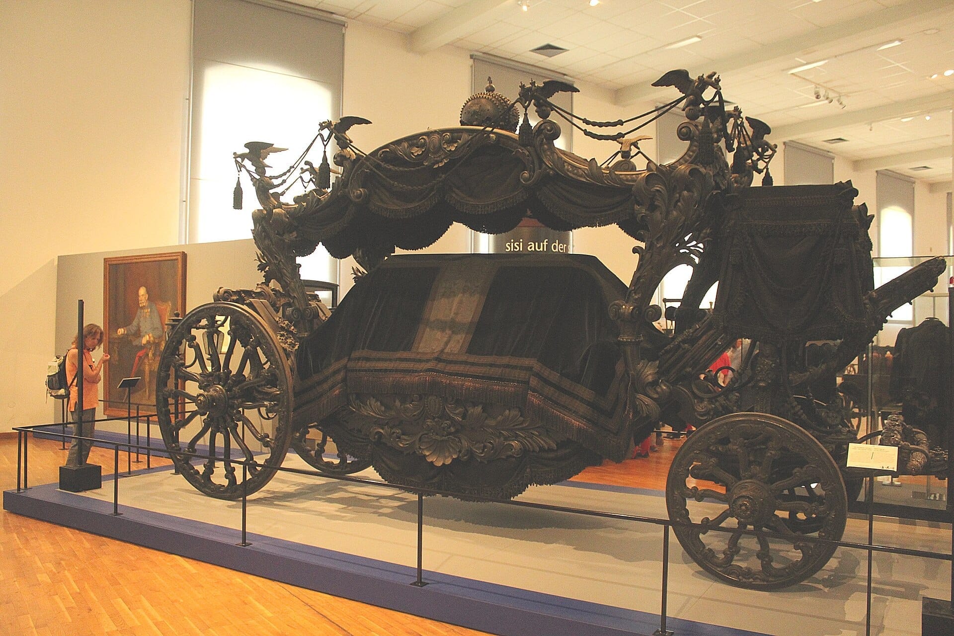 Verzierter schwarzer Leichenwagen ausgestellt im Inneren des Sisi Museum Wien, mit einem Besucher, der Details und Kunstwerke in einem hellen, geräumigen Saal betrachtet.