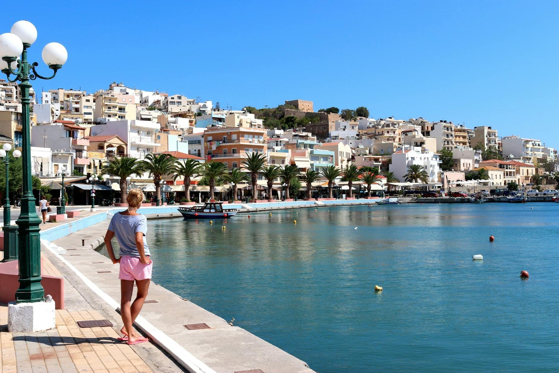 Vista del colorato lungomare di Sitia con palme, barche ormeggiate e case sulla collina sotto un cielo azzurro limpido nella Creta orientale.