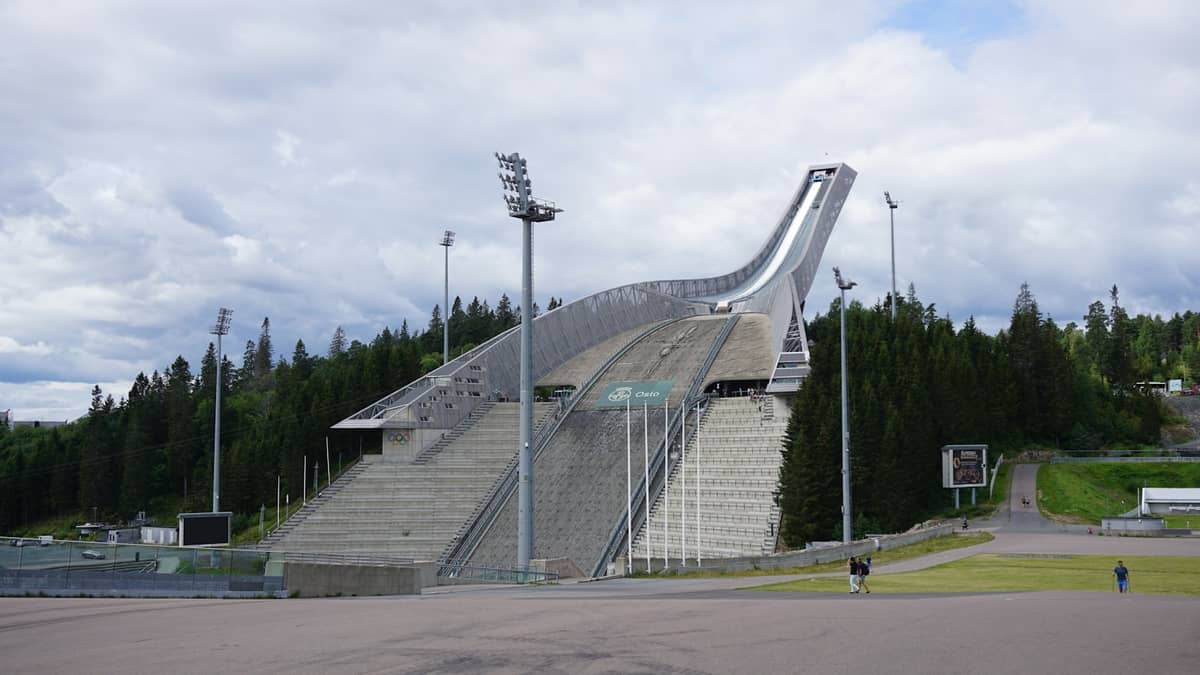 ภาพกว้างของหอคอยสกีจัมป์ Holmenkollen ในออสโลช่วงหน้าร้อน มีโครงสร้างคอนกรีต ป่าล้อมรอบ และผู้คนบางส่วนใกล้ ๆ
