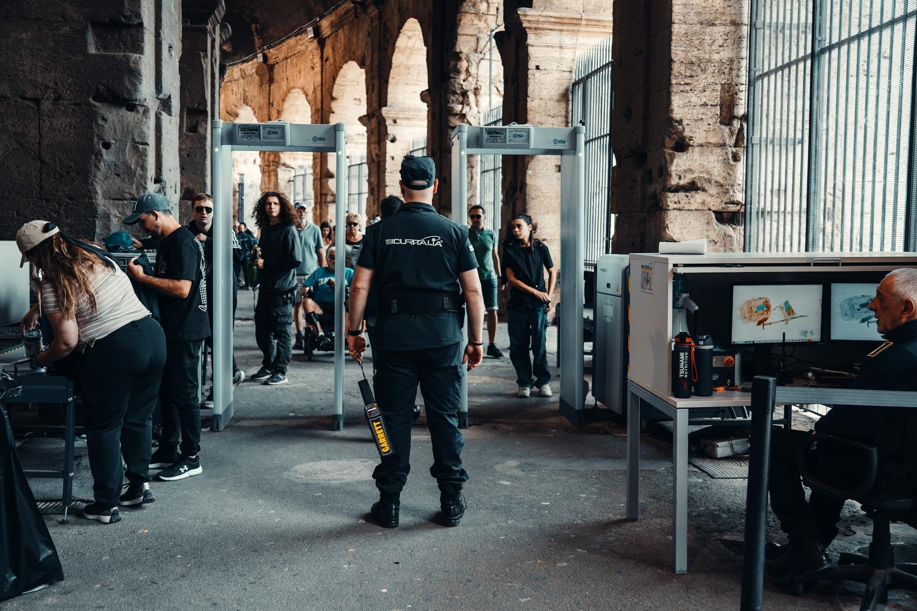 Tourists passing through security screening with metal detectors and x-ray machines inside a Roman landmark, monitored by security personnel.