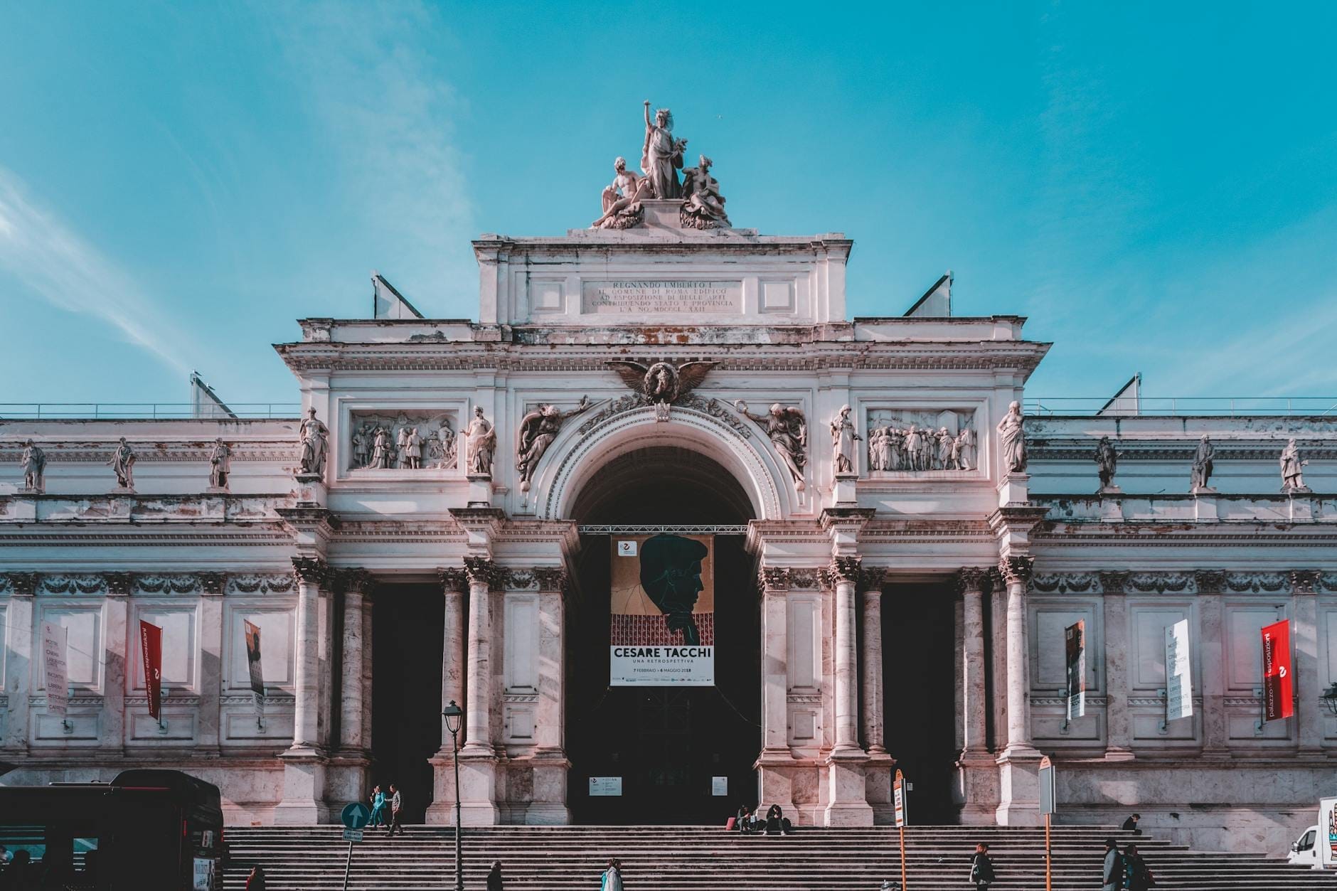 Wide steps and stately facade of a major Rome museum with banners and visitors in front under blue sky.