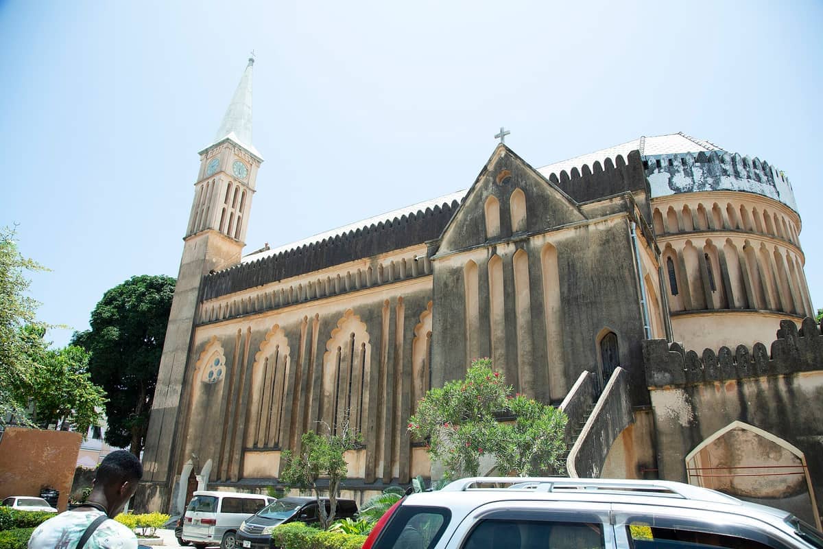 Esterno della Cattedrale Anglicana Christ Church a Stone Town, Zanzibar, con architettura storica, torre dell'orologio e auto parcheggiate alla luce del giorno.