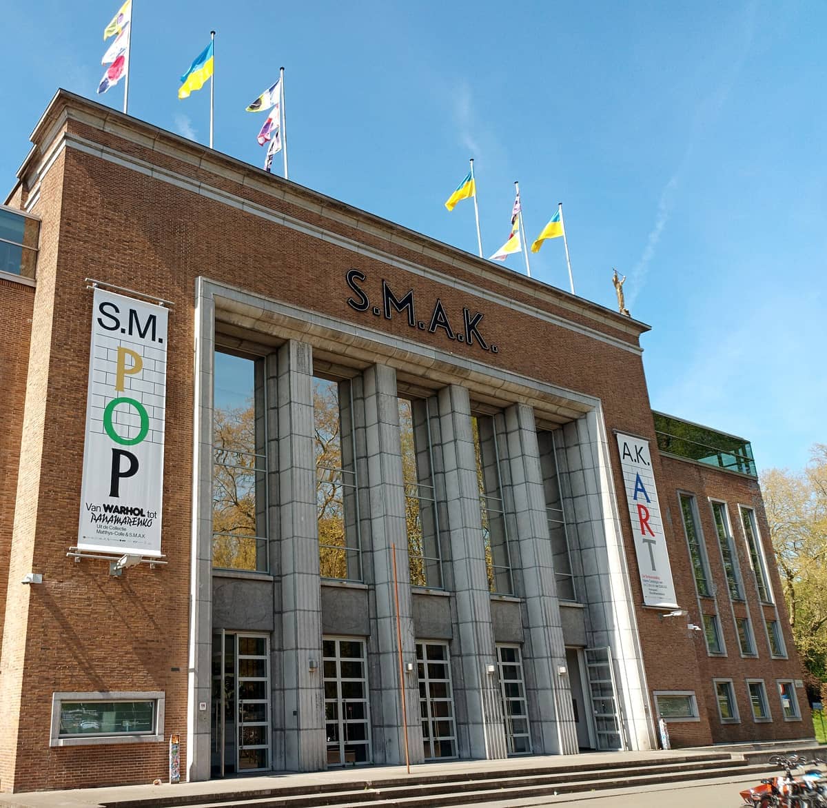 Exterior of S.M.A.K. in Ghent showing the brick facade, tall columns, flags on the roof, and banners promoting current exhibitions.