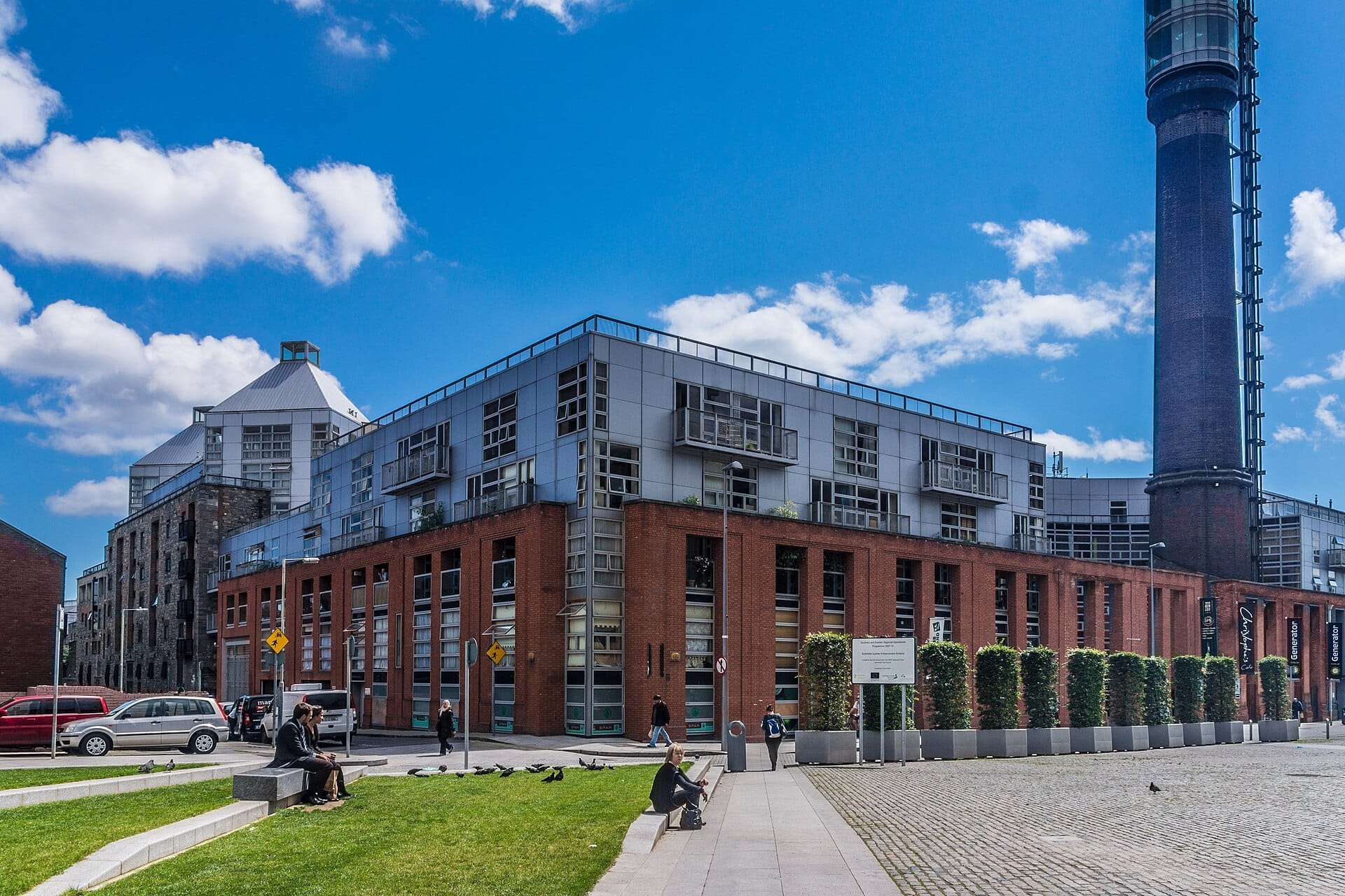 Moderni edifici in mattoni e metallo costeggiano Smithfield Square a Dublino, con persone sedute su panchine erbose sotto un cielo azzurro brillante.