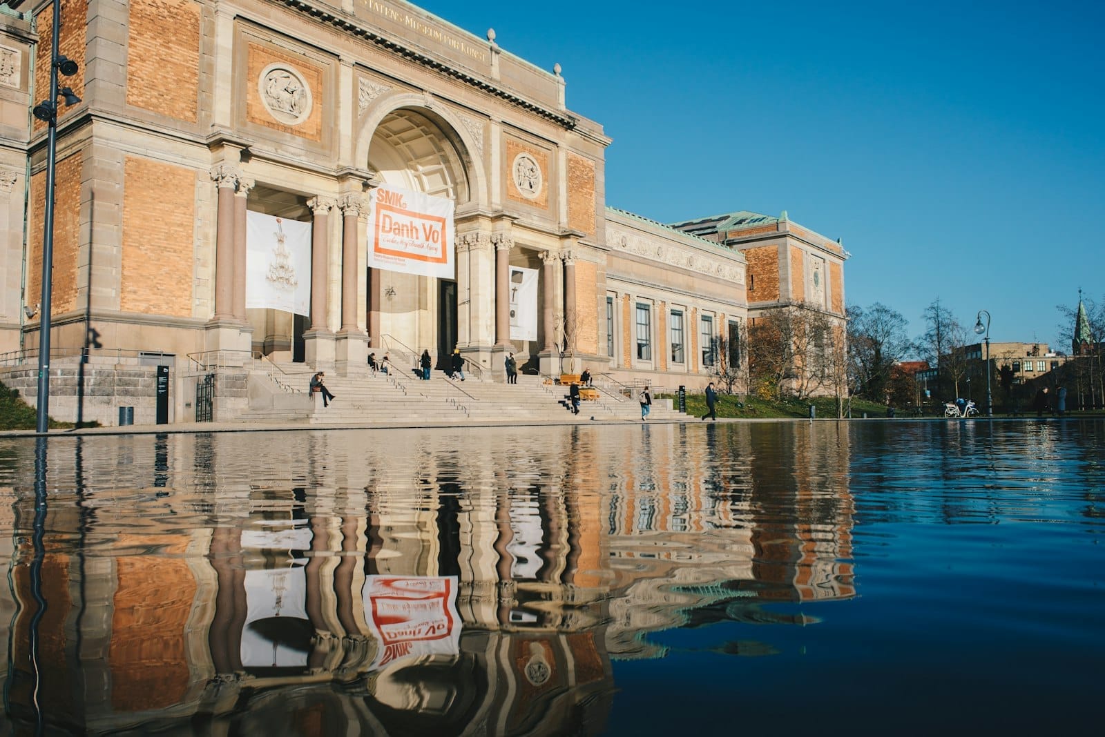 Facciata principale dello SMK – Statens Museum for Kunst di Danimarca, con grande scalinata, vasca riflettente e visitatori sotto un cielo azzurro limpido.