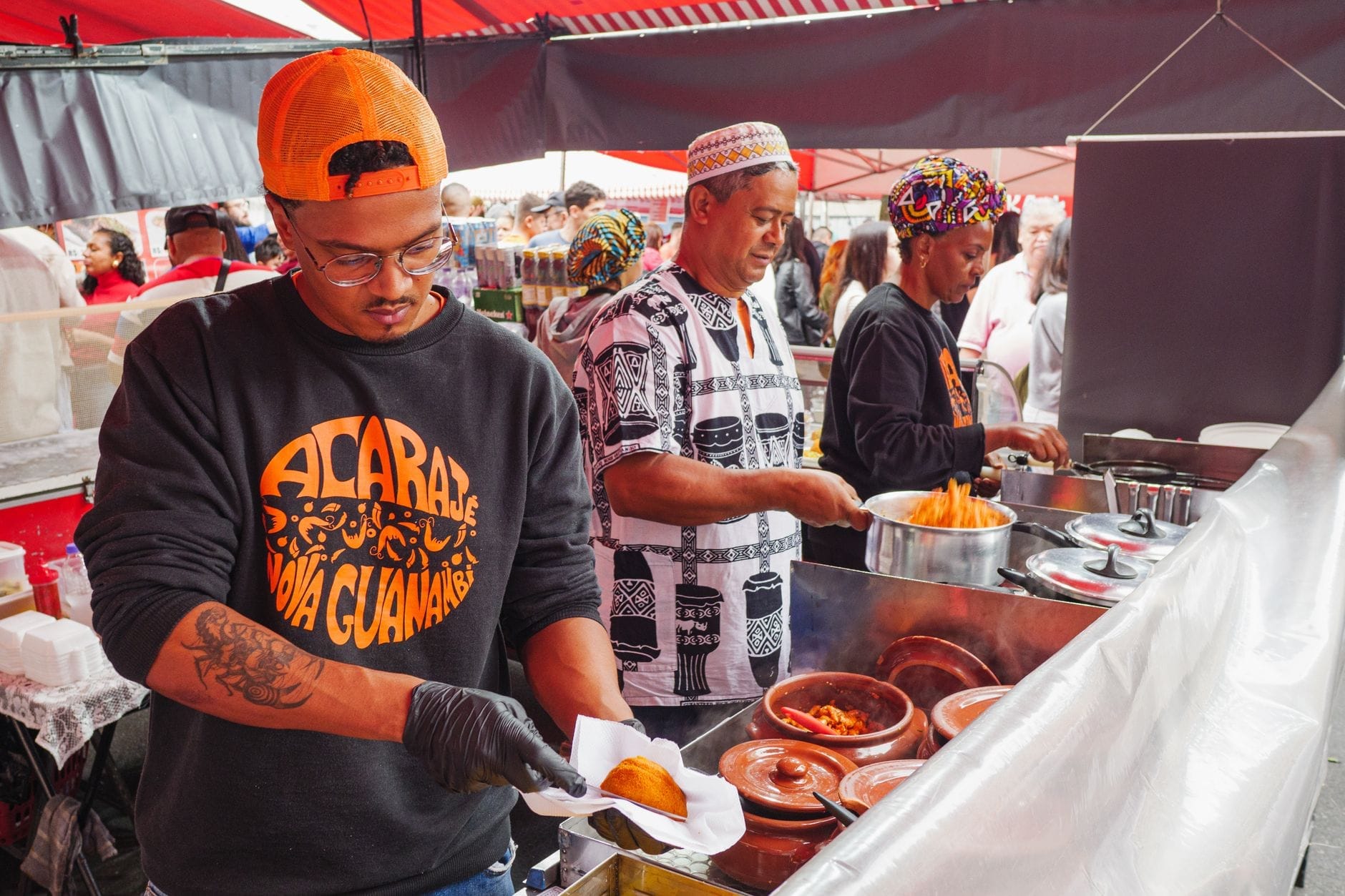 I venditori preparano e servono cibo al loro colorato stand durante lo Smorgasburg, il mercato all'aperto di Brooklyn, sotto tende rosse con una folla sullo sfondo.