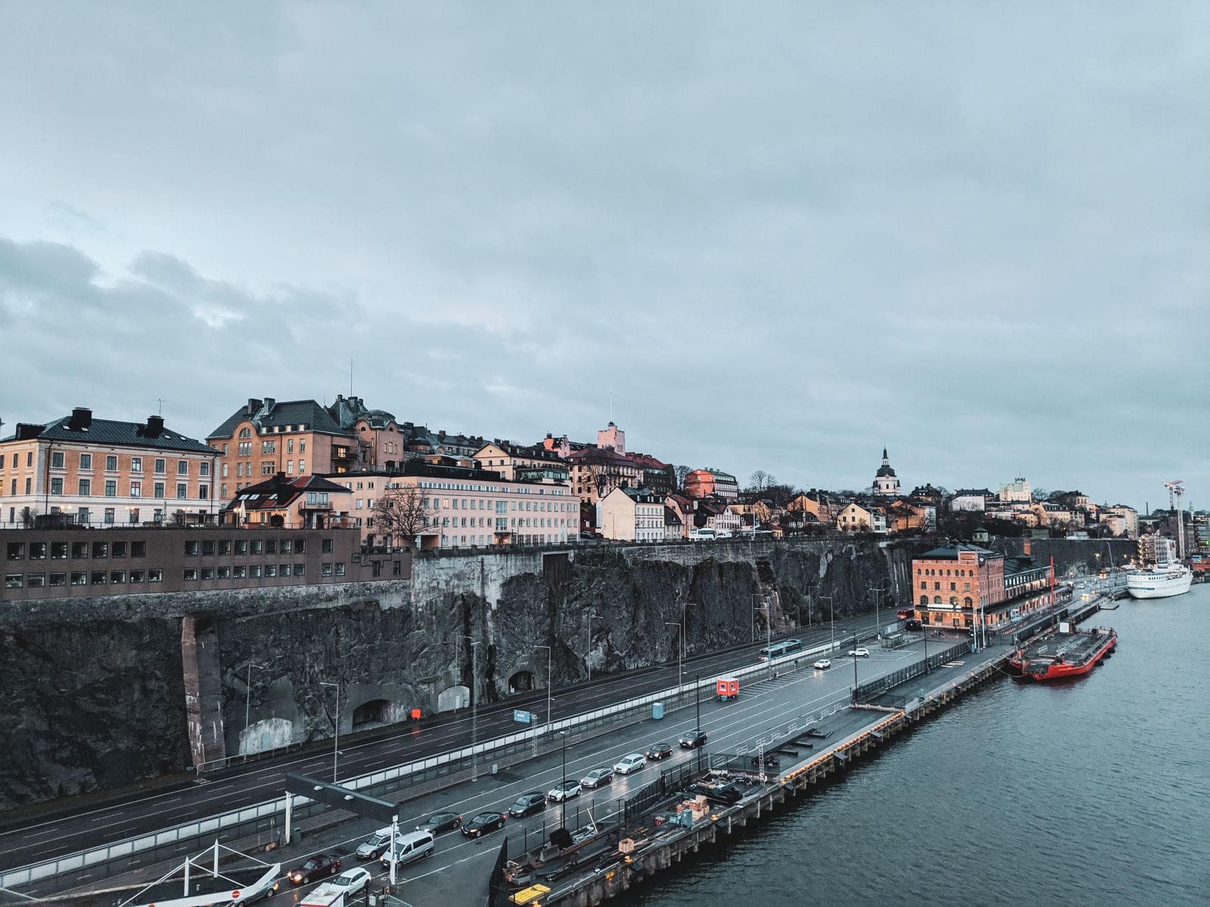 วิวตึกบนหน้าผาเกาะ Södermalm และถนนเลียบชายฝั่ง มีรถกับเรือริมคลอง ใต้ท้องฟ้ามีเมฆ
