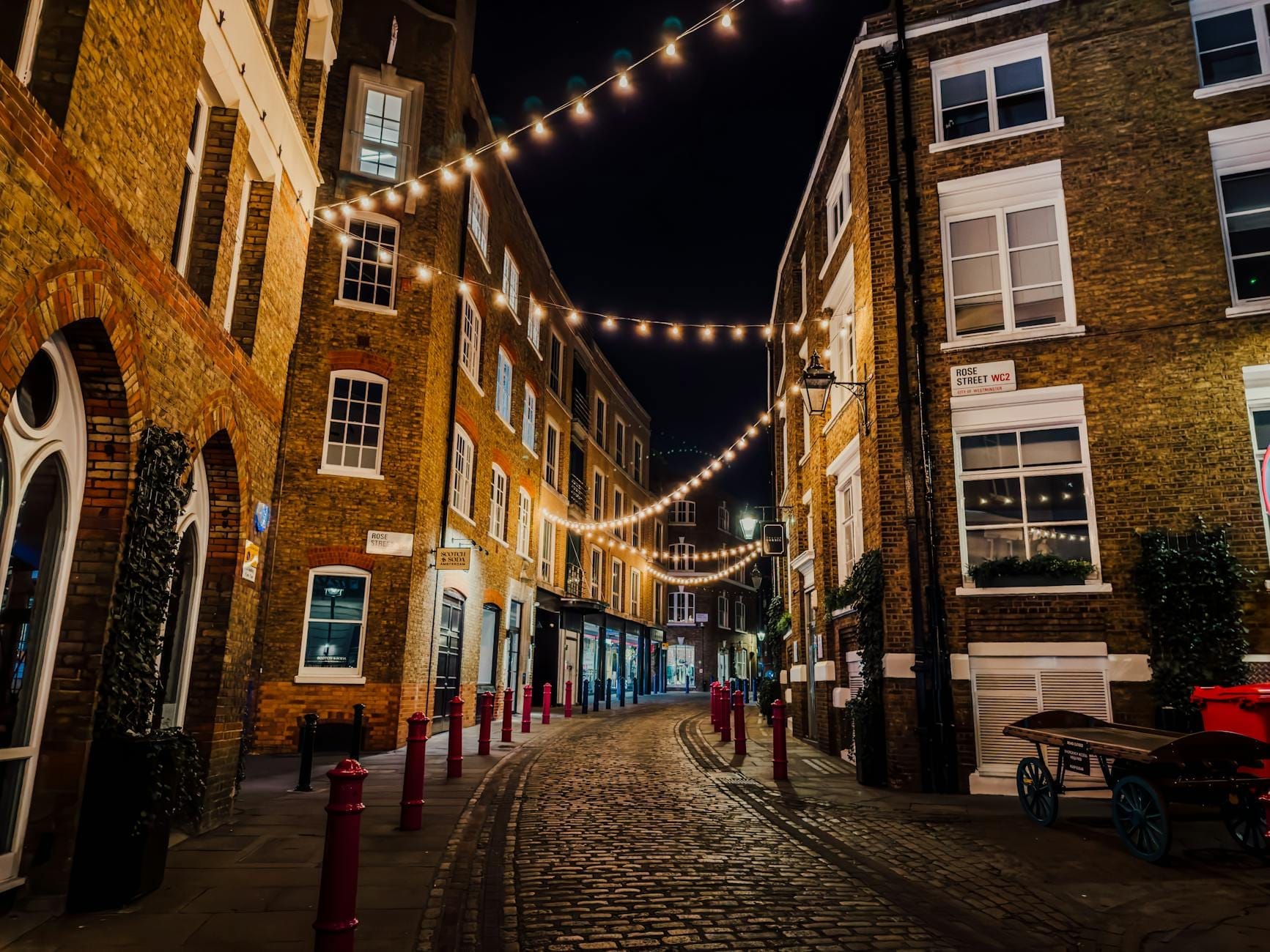 Una caratteristica strada acciottolata di Soho di notte, fiancheggiata da edifici in mattoni e illuminata da calde luci festoni, che evocano l'energia vivace del quartiere dopo il tramonto.