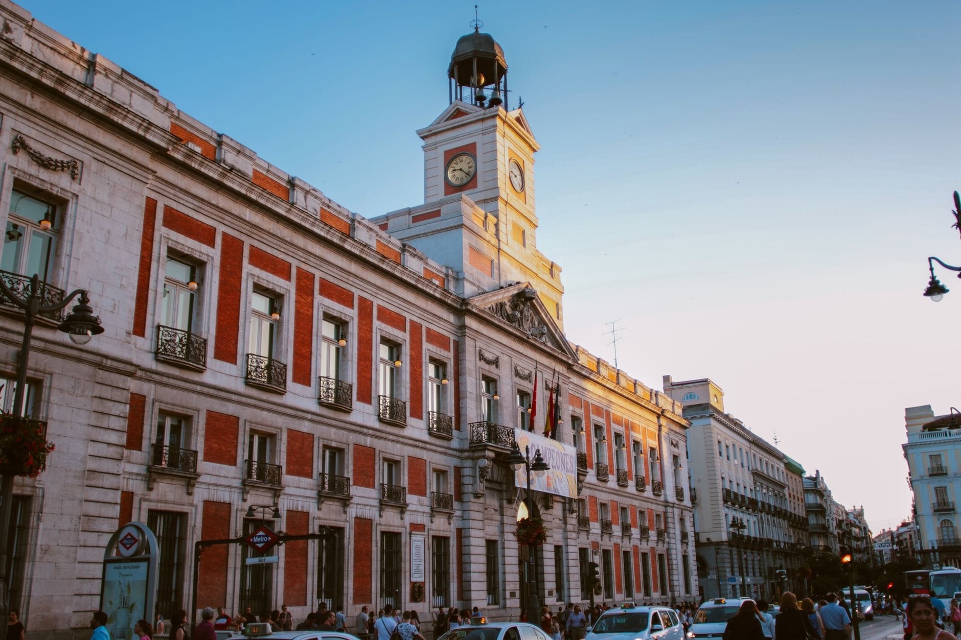 พระอาทิตย์ตกที่ Puerta del Sol เห็นหอนาฬิกาสีแดงและขาว ตึกประวัติศาสตร์ และผู้คนจำนวนมาก สะท้อนพลังใจกลางเมืองของมาดริด