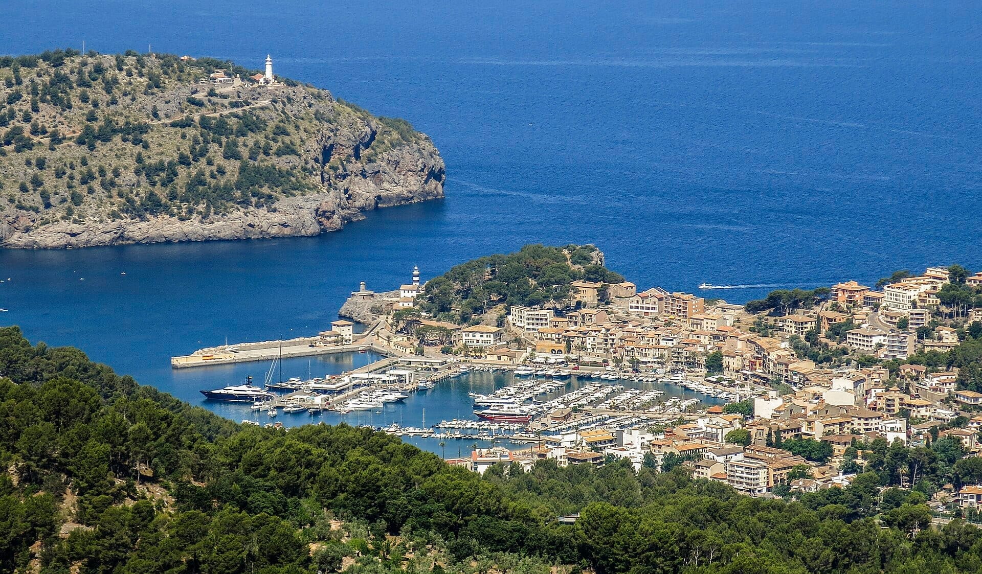 Vista aerea del Port de Sóller con un porto turistico pieno di barche, le acque azzurre del Mediterraneo, colline verdi e un faro storico.