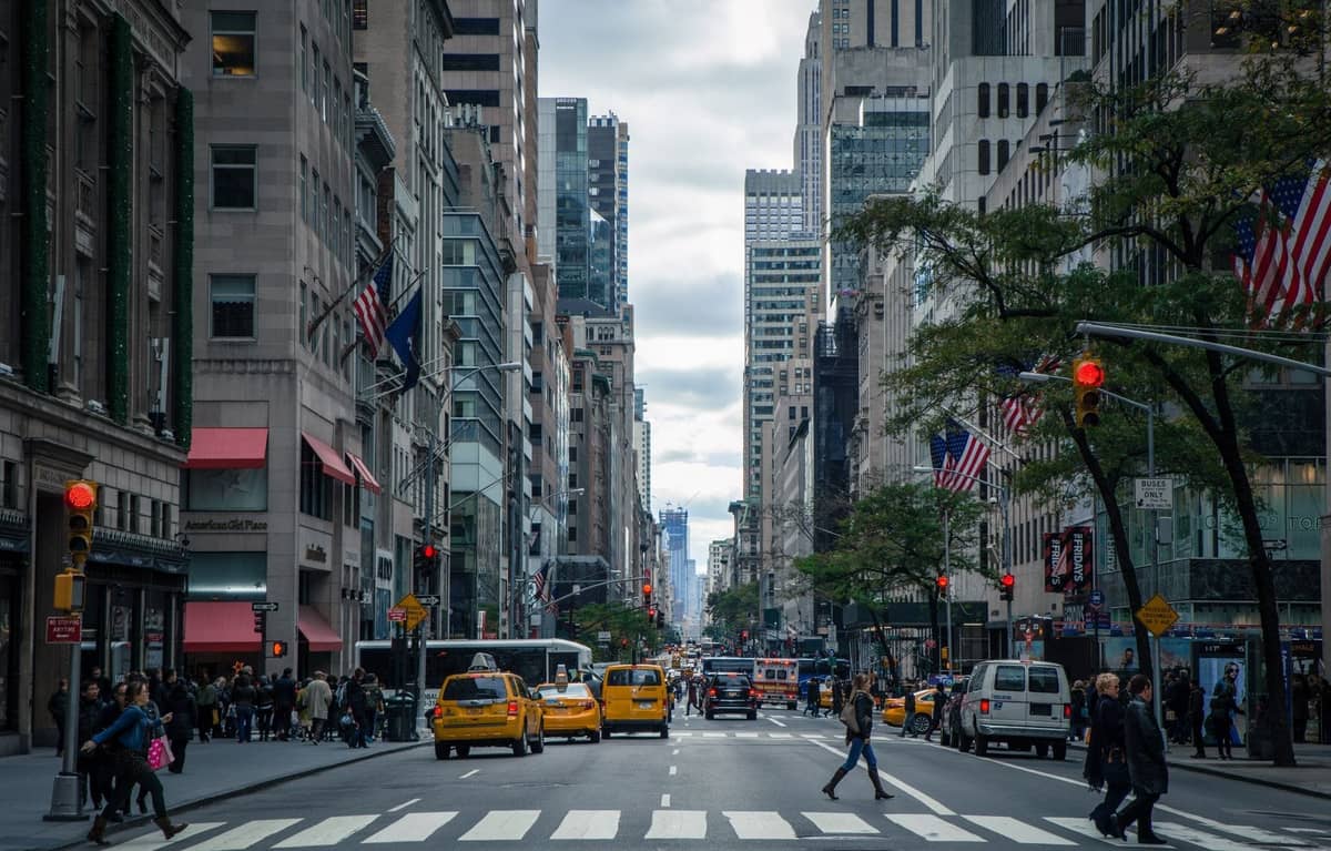 Belebte Straße in New York City mit gelben Taxis, überquerenden Fußgängern und hohen Gebäuden auf beiden Seiten, die die lebhafte Atmosphäre der Stadt einfangen.