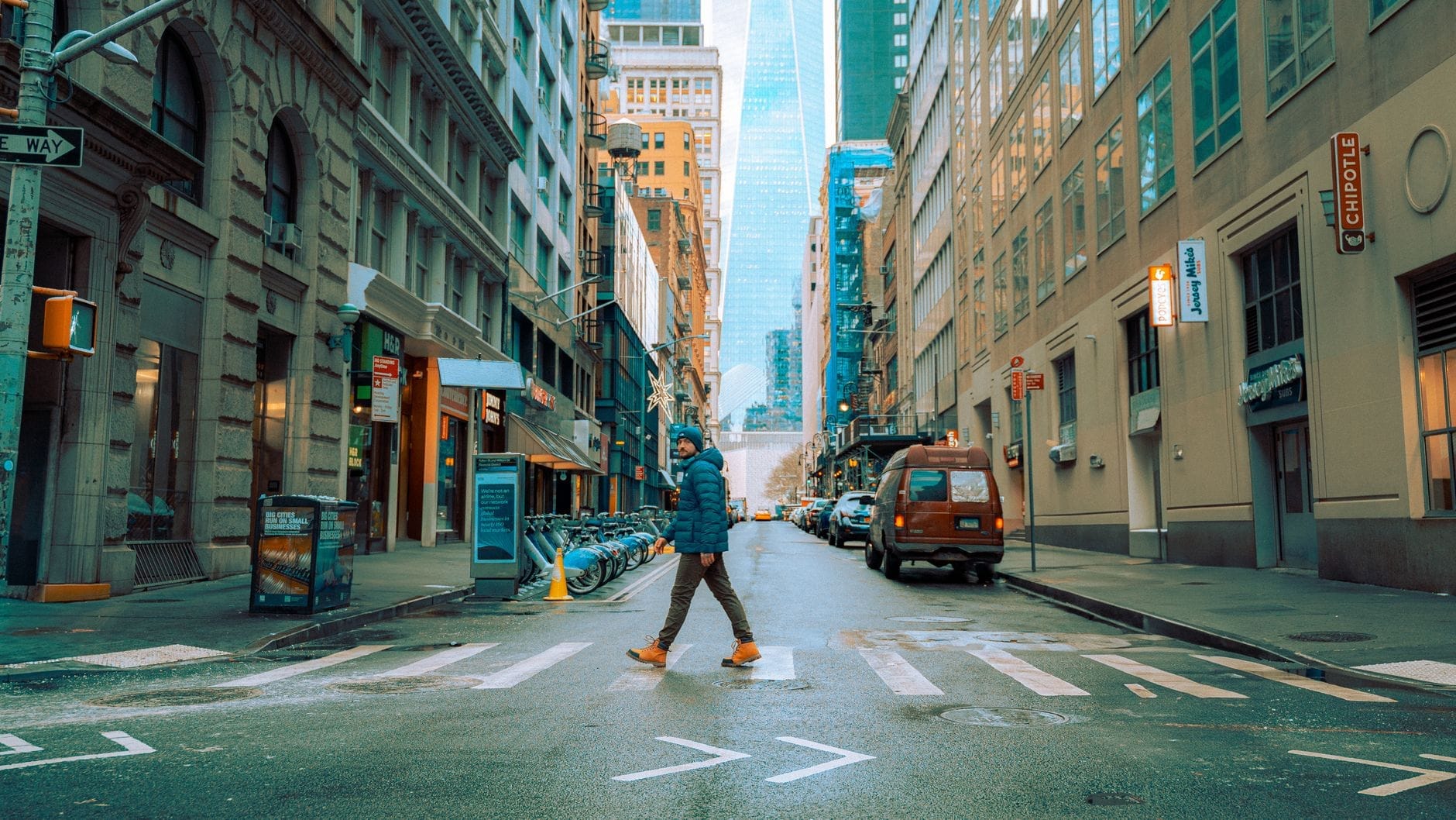 Uma pessoa caminha sozinha por uma faixa de pedestres no meio de uma rua em Manhattan, cercada por arranha-céus e luzes da cidade, evocando a exploração solo em Nova York.