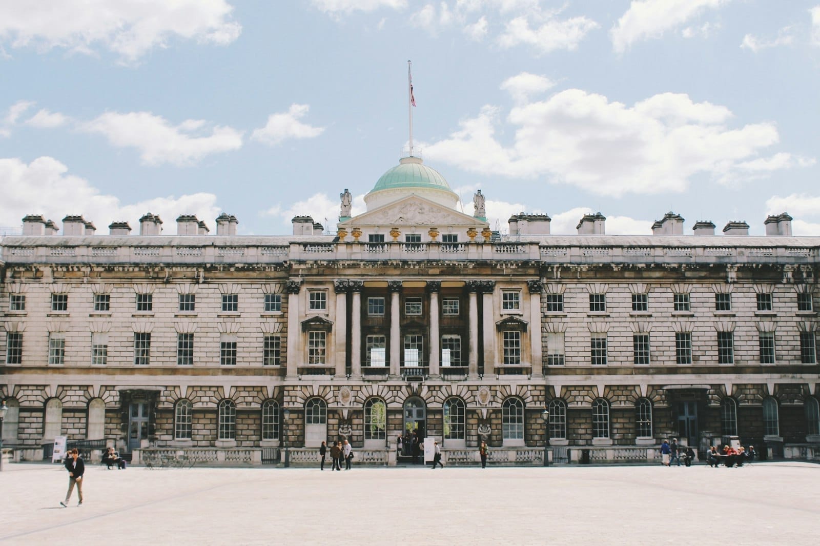 La grandiosa facciata neoclassica di Somerset House con una cupola centrale, colonne in pietra e un ampio cortile sotto un cielo luminoso e parzialmente nuvoloso.