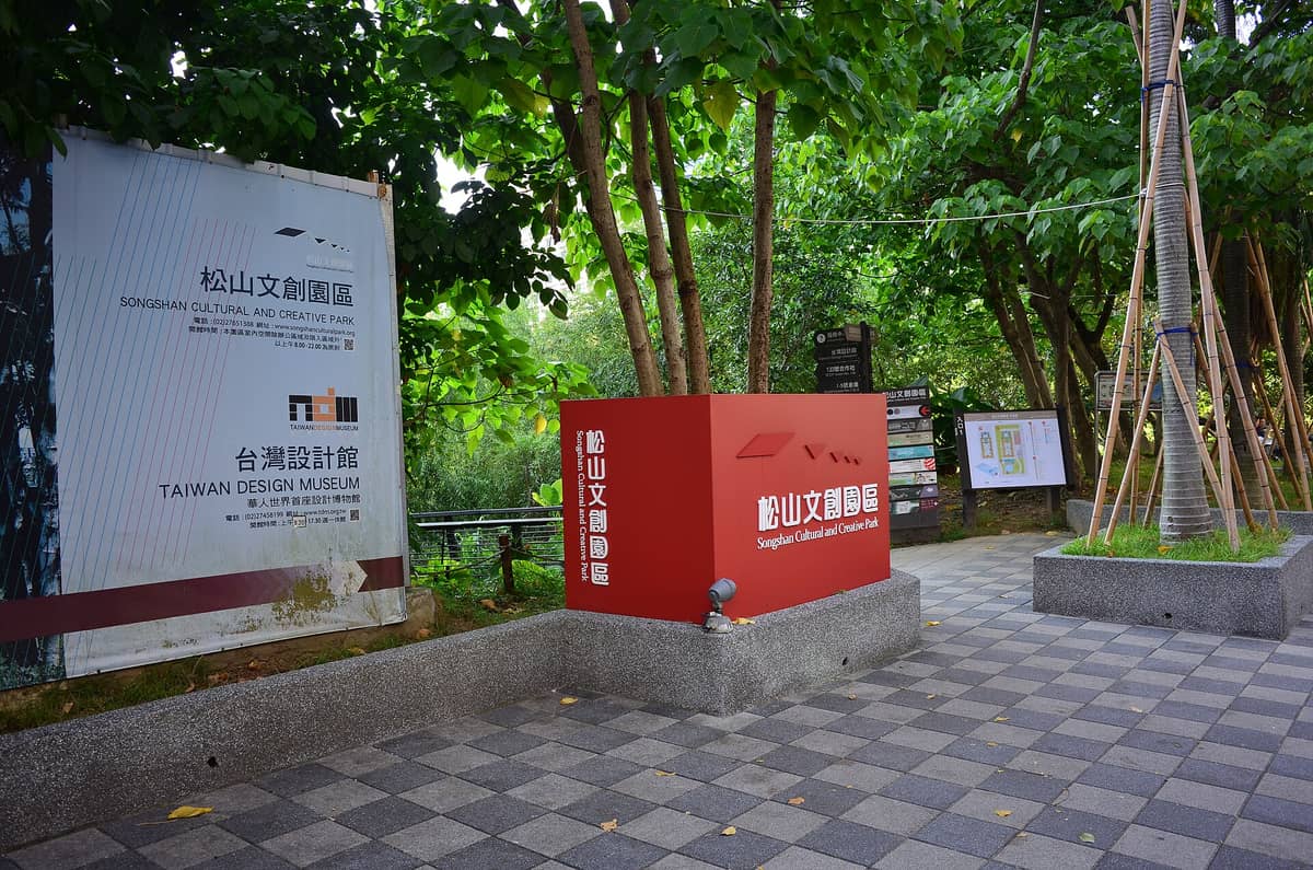 Eingang zum Songshan Cultural and Creative Park mit einem roten Schild und einem großen Banner des Taiwan Design Museum, umgeben von grünen Bäumen.
