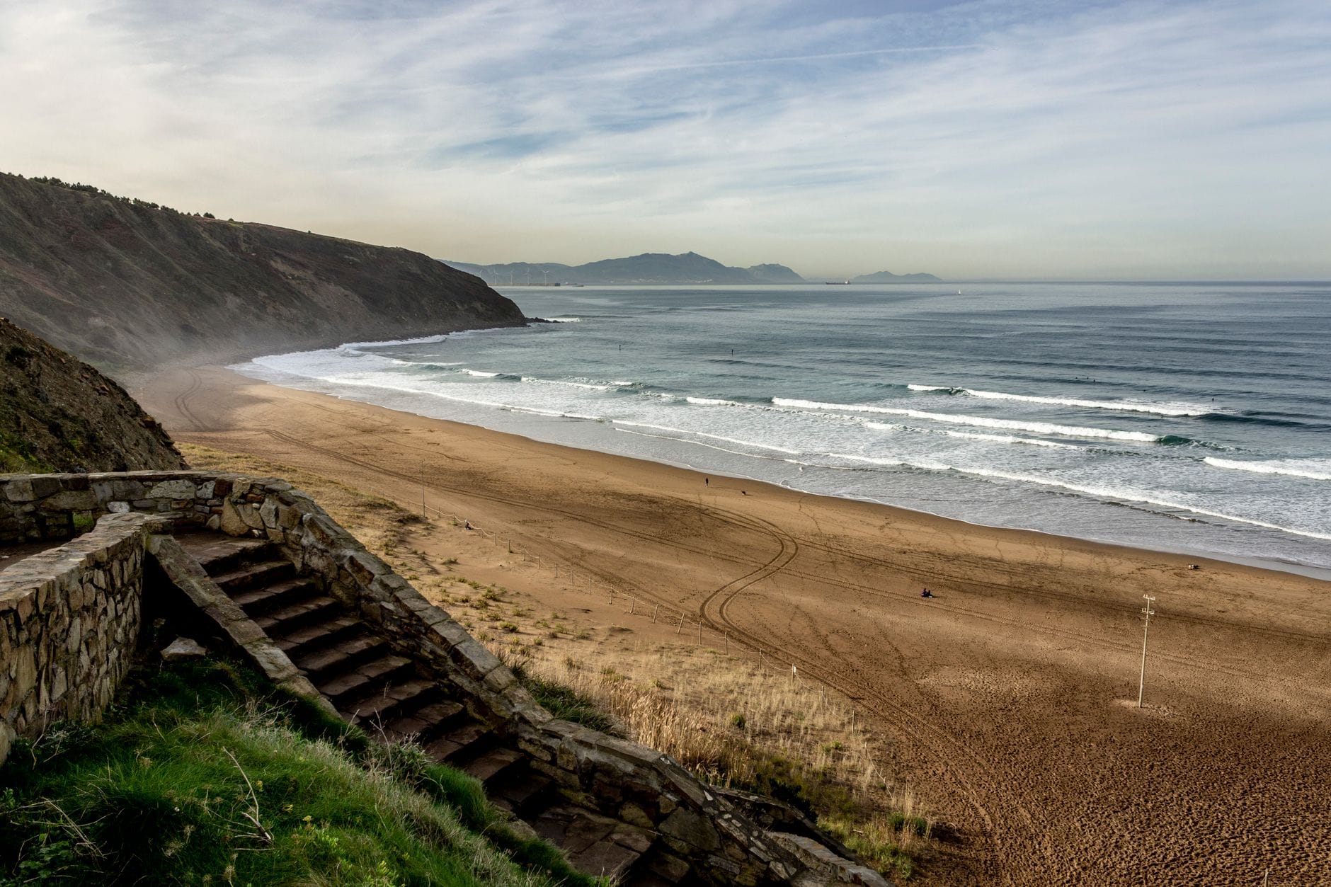 Weiter Blick über den Strand Sopelana mit goldenem Sand, rollenden Wellen, malerischen Klippen und Steinstufen zum Strand hinunter – bei leicht bewölktem Himmel.