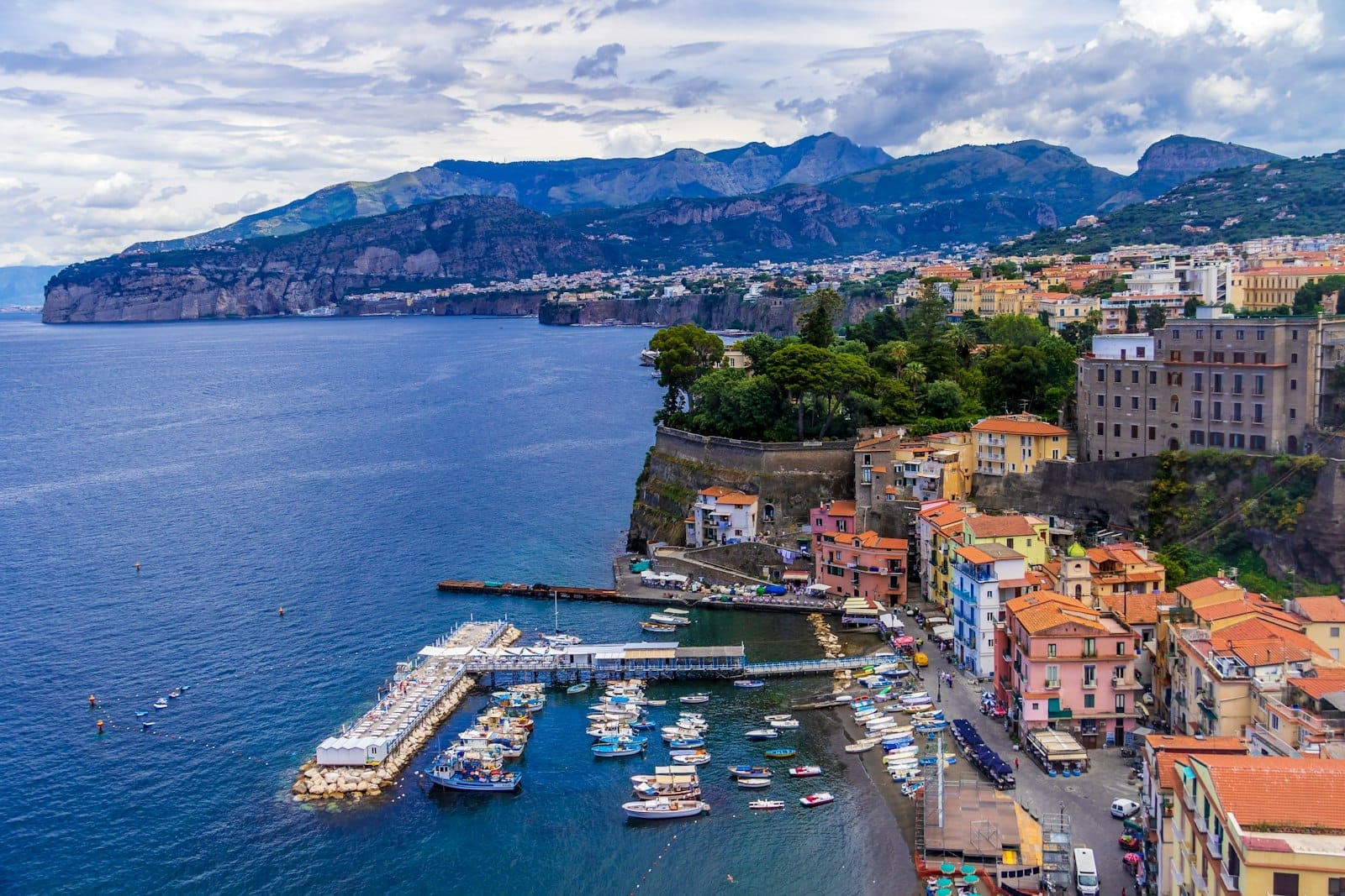 Vista aerea degli edifici colorati sul lungomare di Sorrento, piccole imbarcazioni ormeggiate nel porto turistico e scogliere frastagliate sopra il Golfo di Napoli sotto cieli nuvolosi.