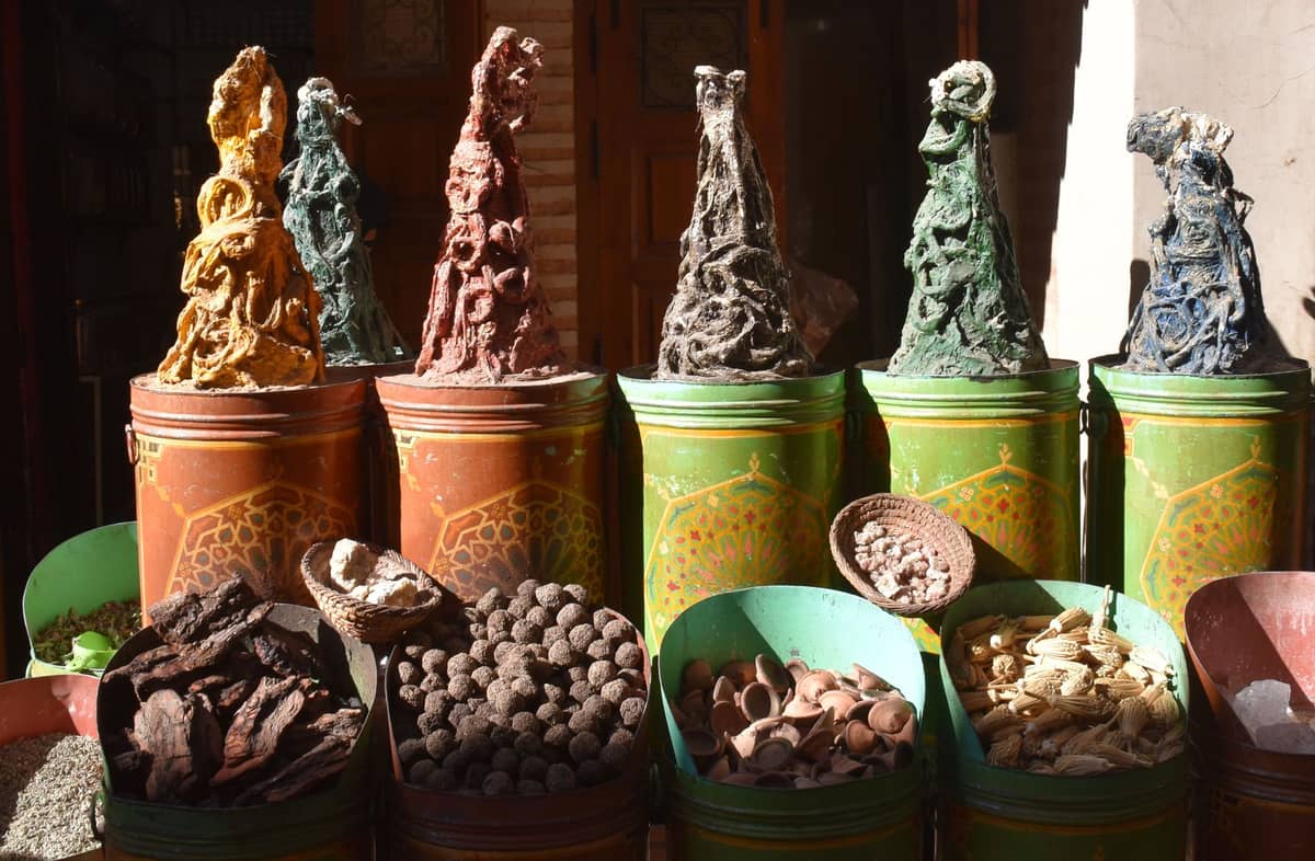 ไหมพรมสีสดแขวนเรียงรายและวัตถุดิบย้อมสีในถาดโลหะที่แผงซูก เดส์ เต็งตูริเยร์ มาราเกช มีแสงแดดลอดเข้ามาเล่นเงา