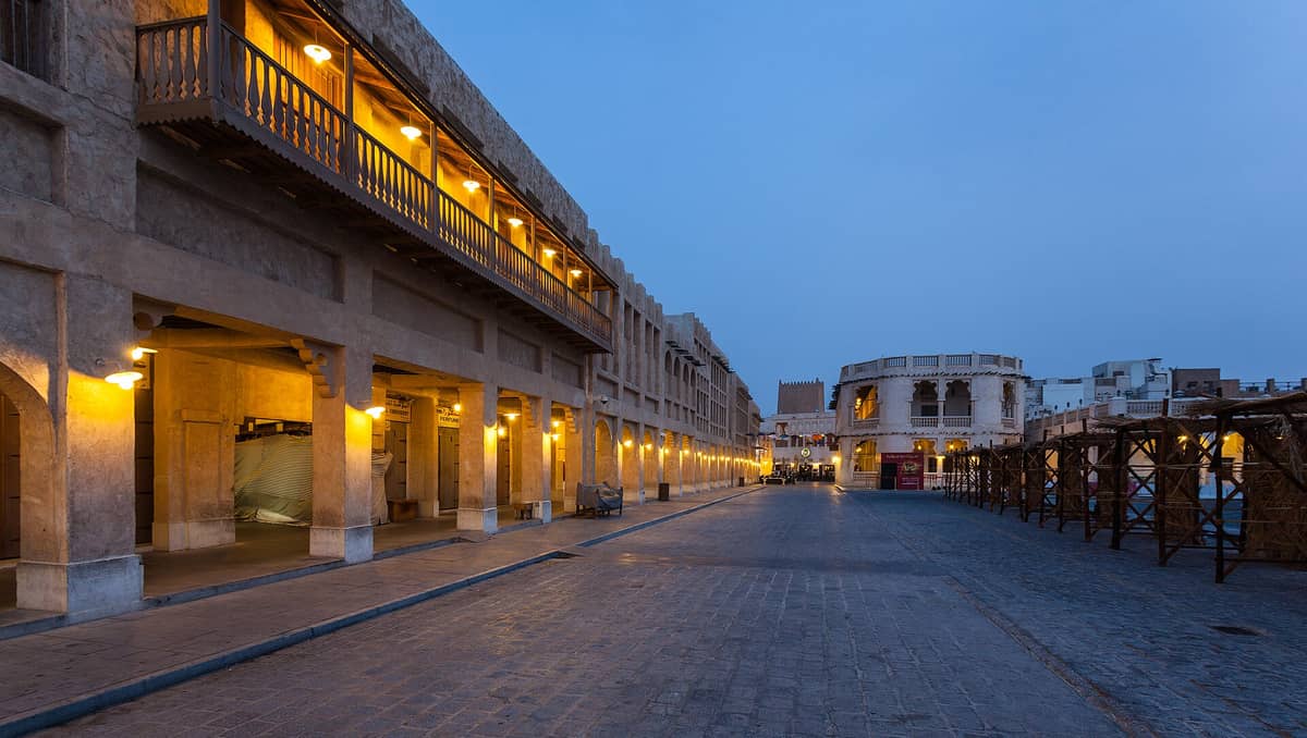 Weitwinkelperspektive auf die Fußgängerhauptstraße des Souq Waqif in der Abenddämmerung: restaurierte Lehmputzfassaden, erleuchtet von Laternen, gesäumt von traditionellen Balkonen.