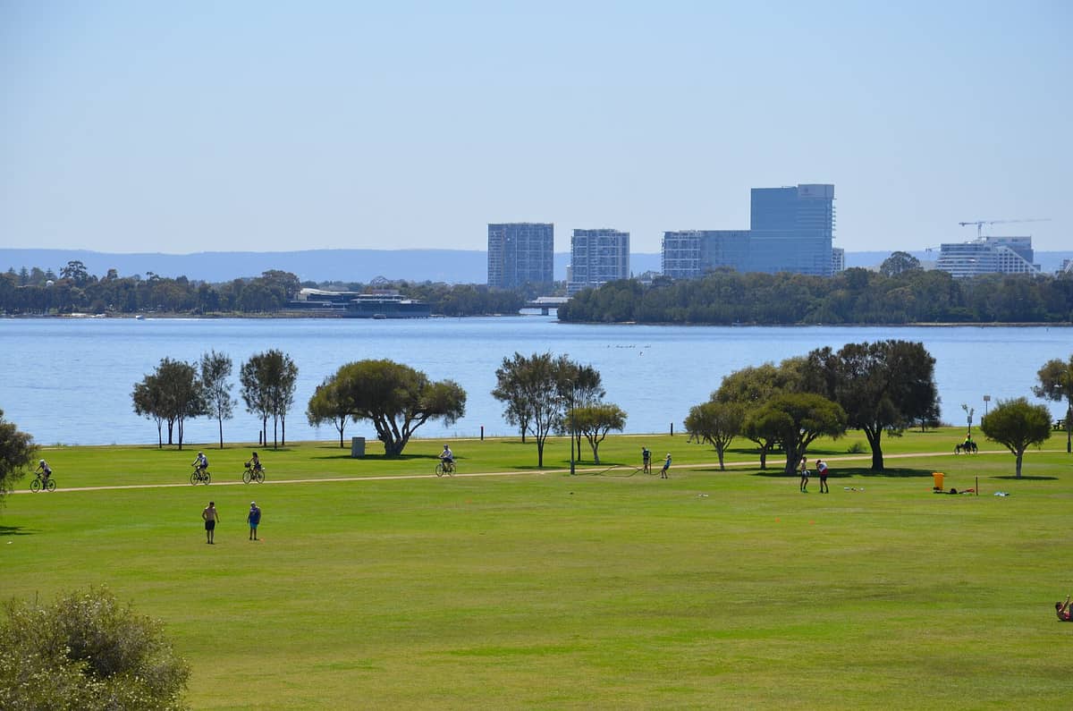 Vista amplia del parque a lo largo de la costanera de South Perth con ciclistas, gente en el césped y el río Swan de fondo bajo un cielo despejado.