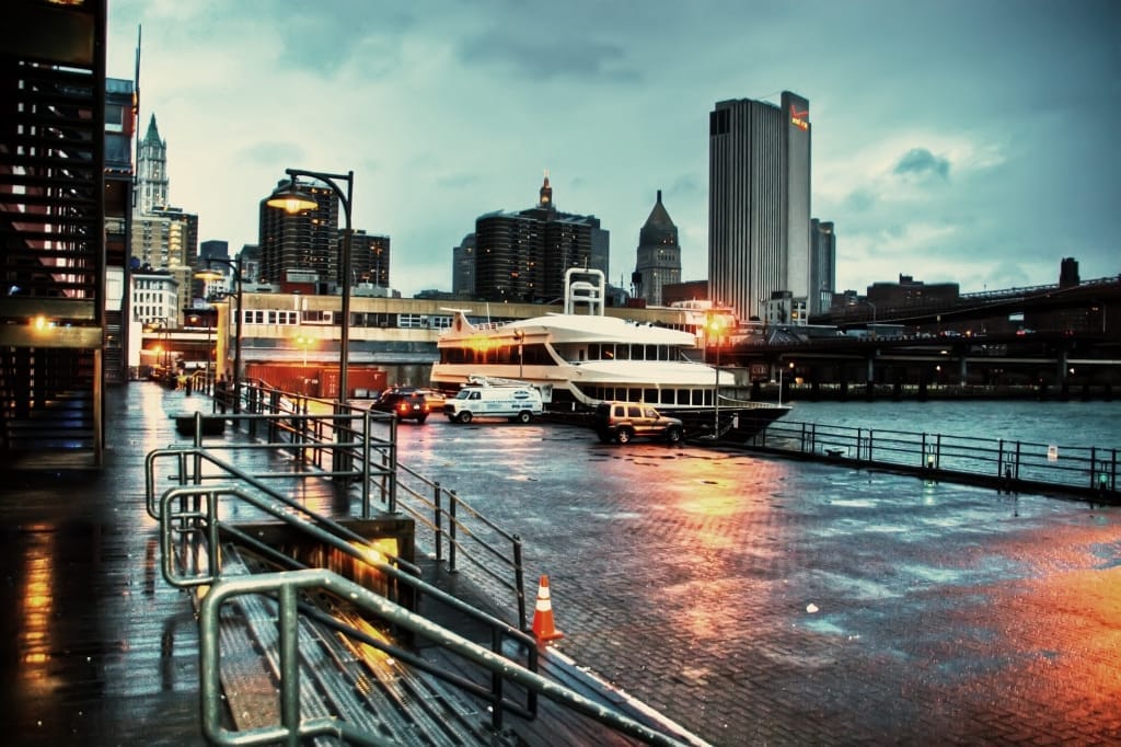 Un molo di ciottoli bagnati nel South Street Seaport Historic District, con una barca attraccata, ringhiere sul lungomare e i grattacieli di Manhattan nella luce serale.
