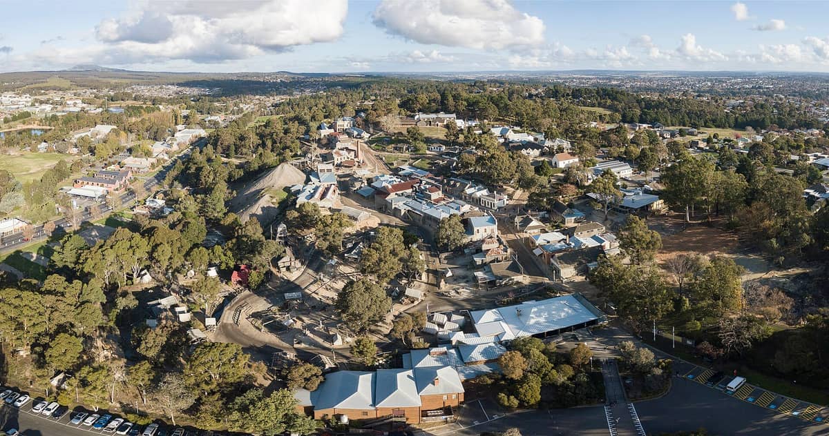 Vista aérea de Sovereign Hill en Ballarat, con edificios históricos, caminos de tierra, árboles y estructuras de minería de oro al estilo de la década de 1850 bajo un cielo soleado.