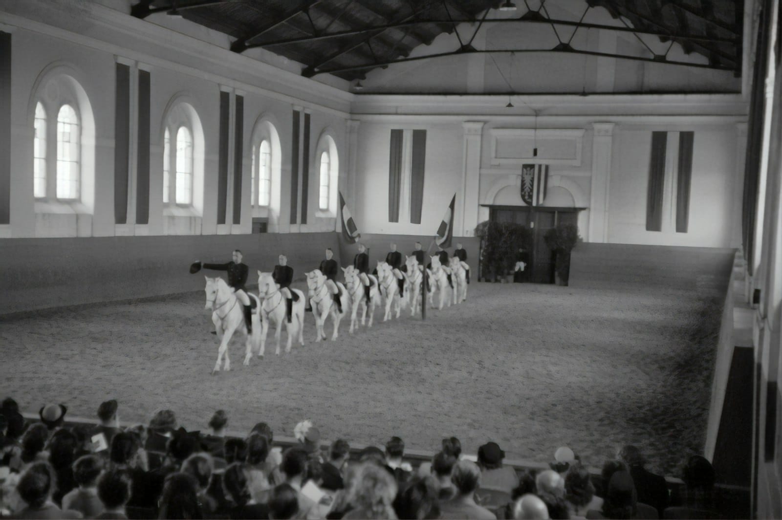 Eine Reihe weißer Lipizzaner mit Reitern in Formation in der barocken Reithalle der Spanischen Hofreitschule, beobachtet von einem sitzenden Publikum.