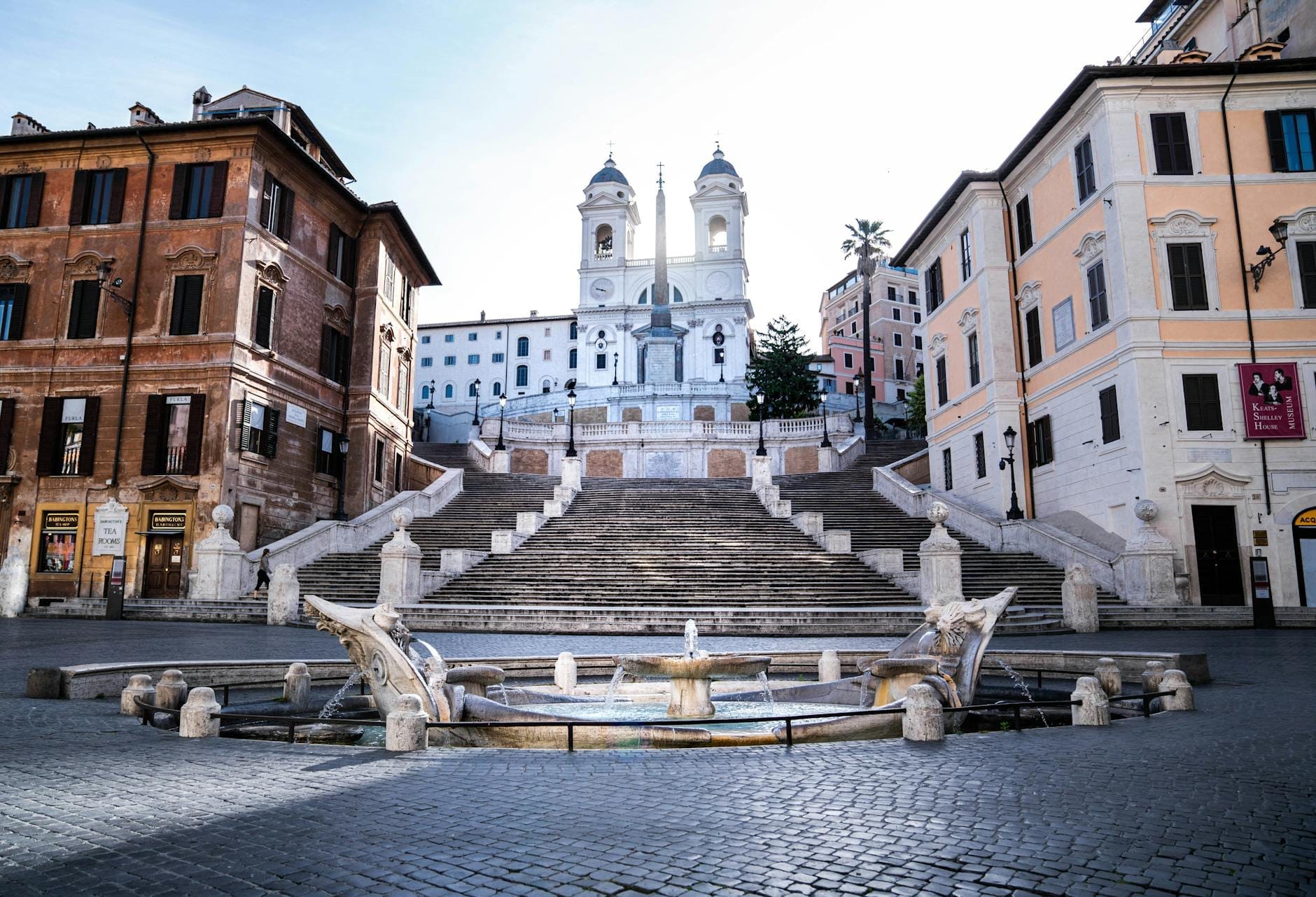 Wide, sunlit view of the Spanish Steps leading up to the Trinità dei Monti church in Rome, with the Barcaccia Fountain and empty piazza below.