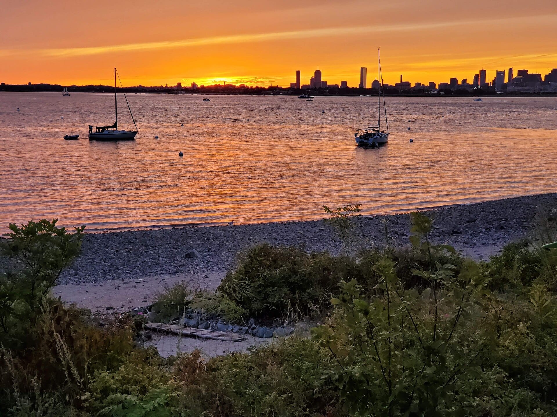Spiaggia di Spectacle Island con barche a vela su acque calme, lo skyline di Boston sullo sfondo e un vibrante tramonto arancione che illumina il cielo.