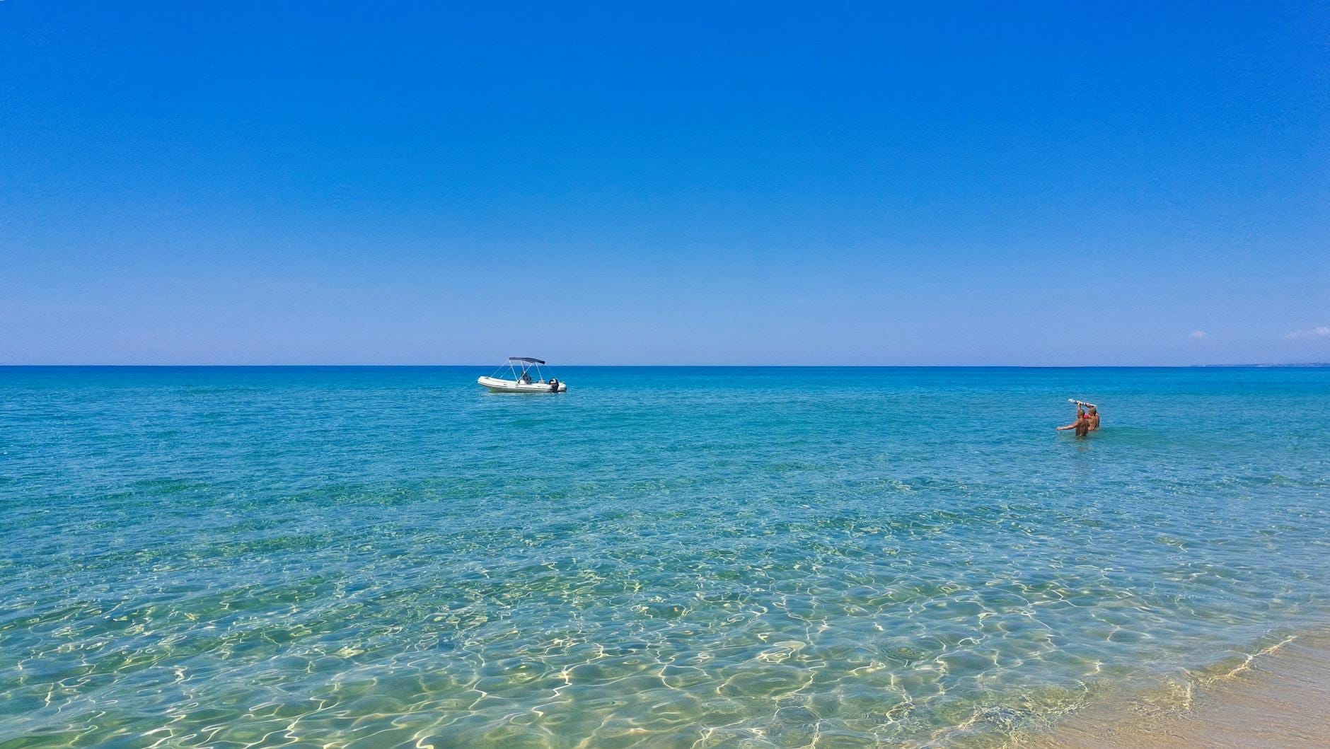 Weiter Blick auf einen ruhigen Strand mit klarem türkisfarbenem Wasser, einem kleinen Boot und einem Schwimmer unter strahlend blauem Himmel.