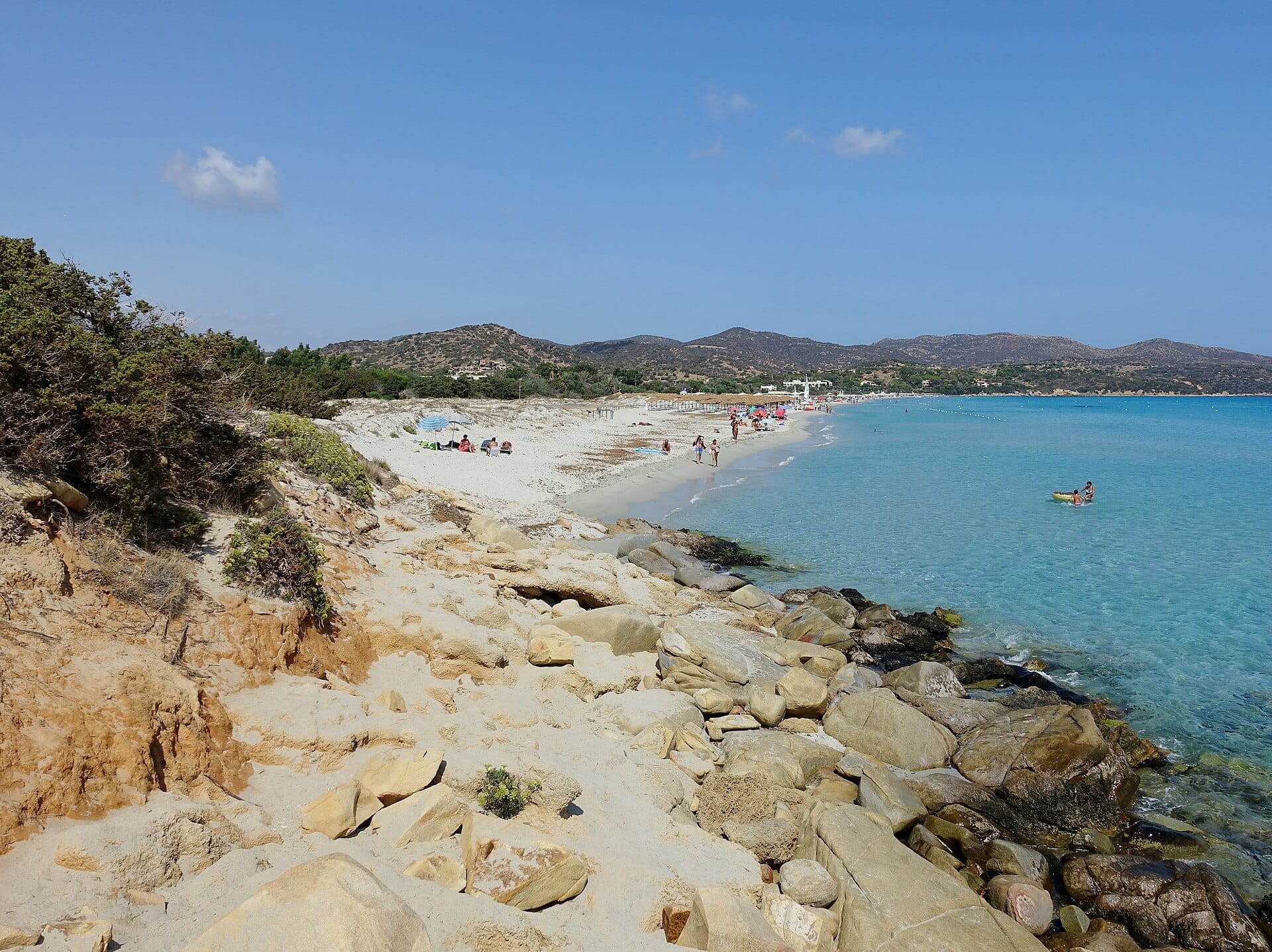 Die Spiaggia di Simius bietet weißen, feinen Sand, türkisfarbenes Wasser, eine felsige Küstenlinie, Sonnenbader und Hügel unter einem klaren blauen Himmel auf Sardinien.