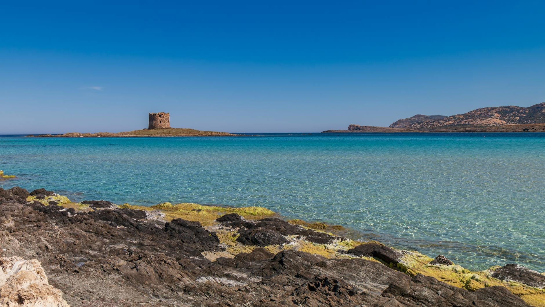 Klares türkisfarbenes Wasser und weißer Sand am Strand La Pelosa in Sardinien, mit einem historischen spanischen Wachturm und Bergen unter strahlend blauem Himmel.