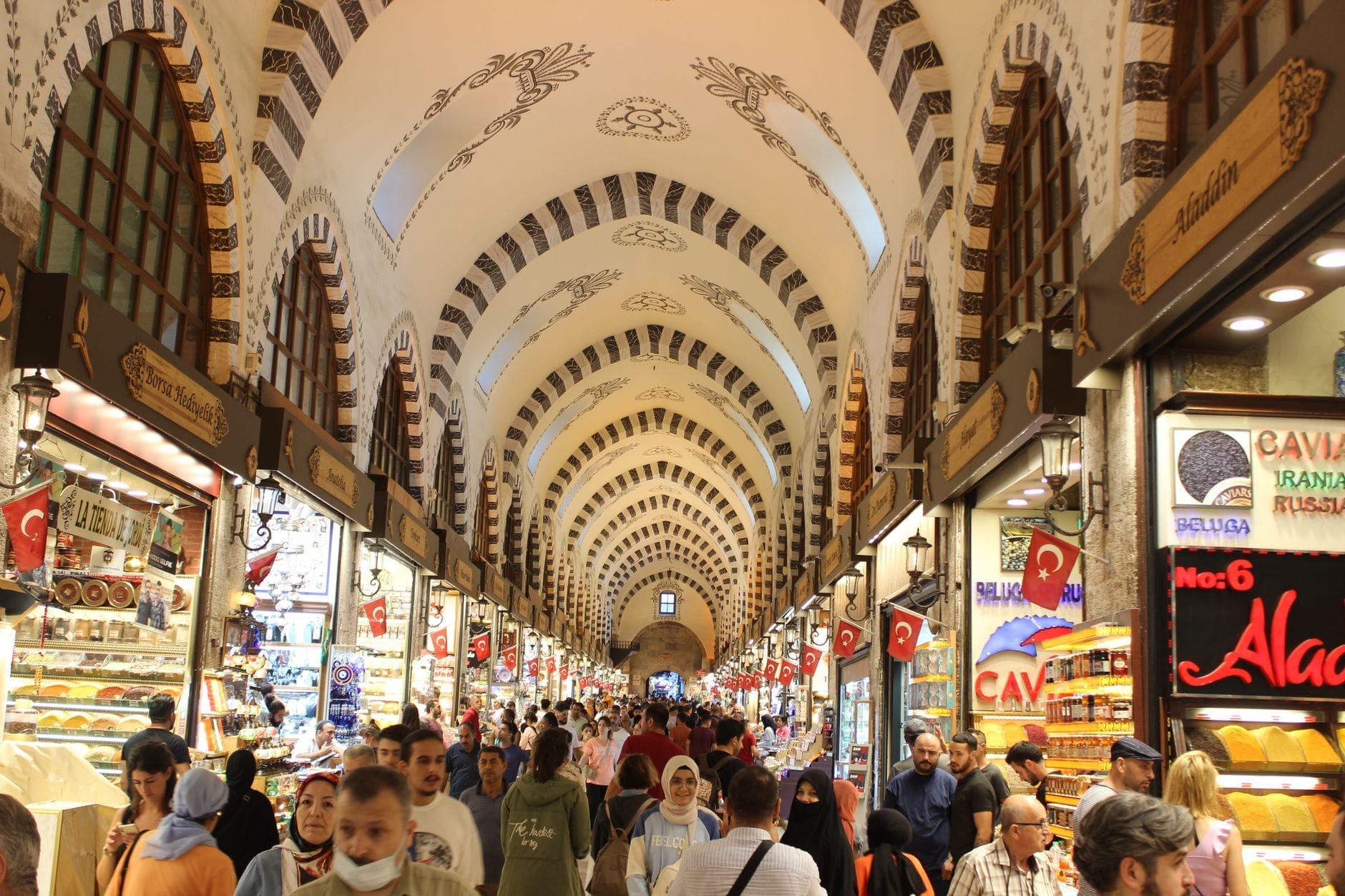 Interno affollato del Bazar delle Spezie di Istanbul, con soffitto ad arco riccamente decorato, visitatori in movimento e colorati negozi di spezie, dolci e lokum su entrambi i lati.