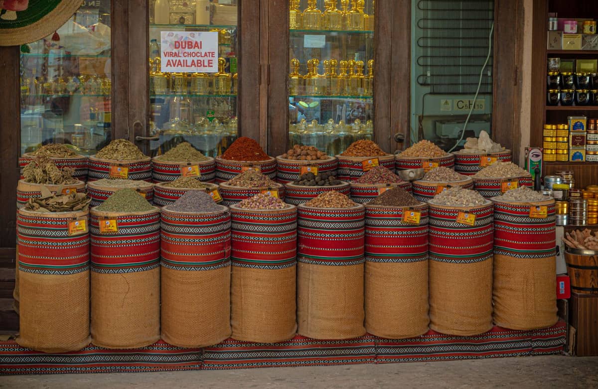 Ein Ladeneingang im Dubais Gewürzsouk mit großen Jutesäcken voller bunter Gewürze, aufgestapelt vor traditionellen Holztüren und Golddekorationen.