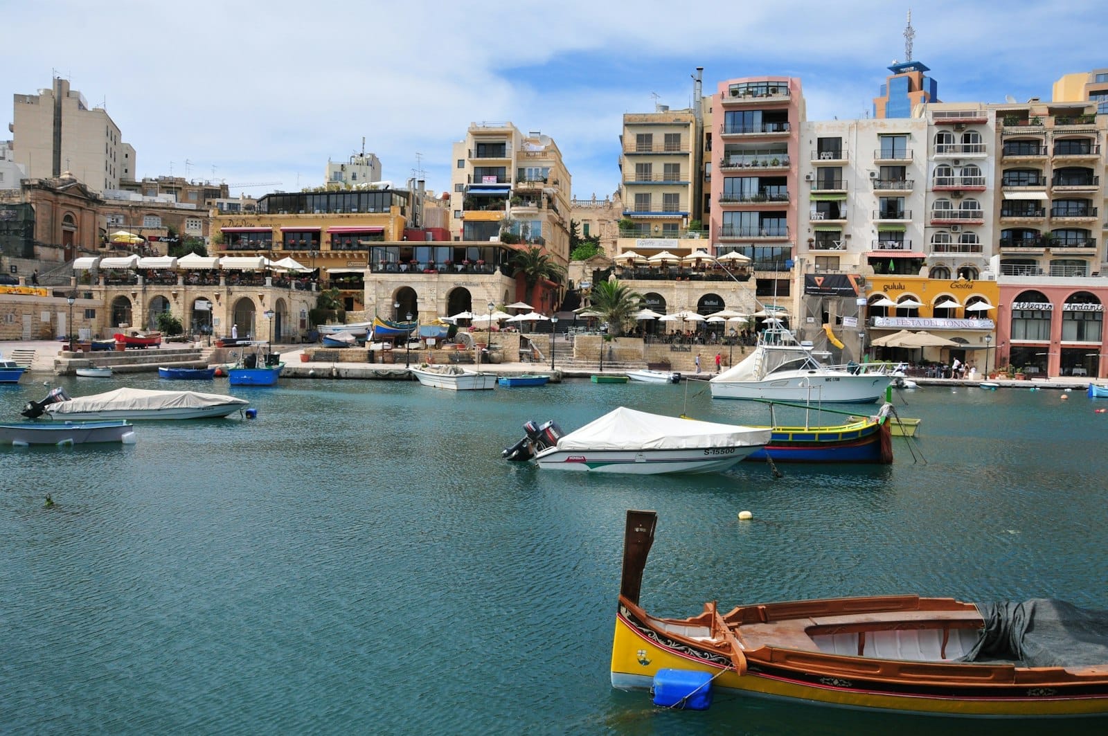 Tradizionali barche da pesca galleggiano nella baia di Spinola, Malta, con edifici colorati, terrazze di ristoranti e acqua blu sotto un cielo parzialmente nuvoloso.