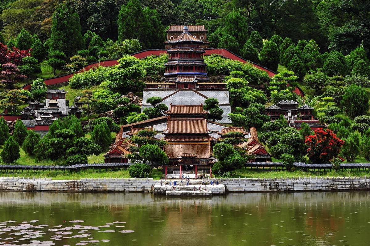 Uma réplica em miniatura de um palácio chinês tradicional rodeado de vegetação exuberante e árvores em Splendid China & China Folk Culture Villages, refletido em um lago.