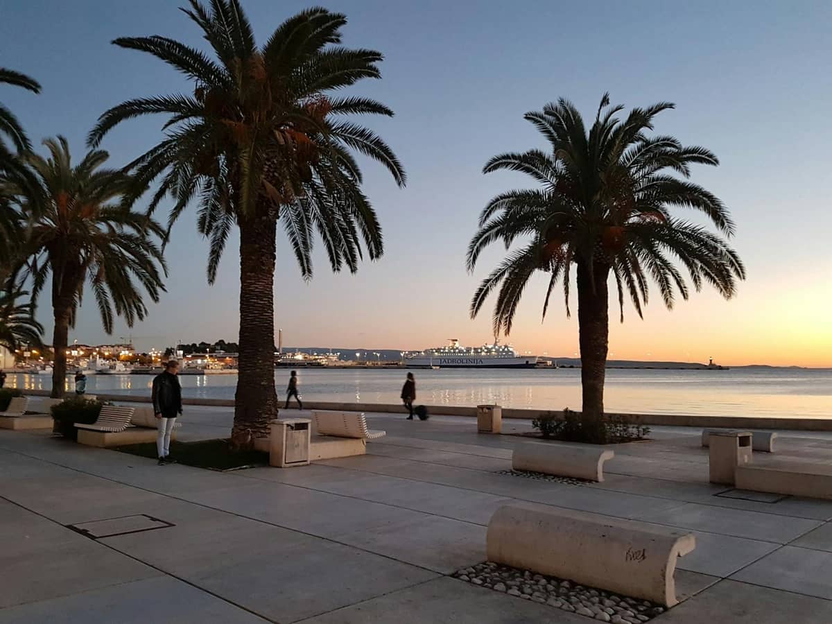 Palmen und Bänke säumen die Riva-Promenade in Split bei Sonnenuntergang, mit Spaziergängern und dem Hafen im Hintergrund.