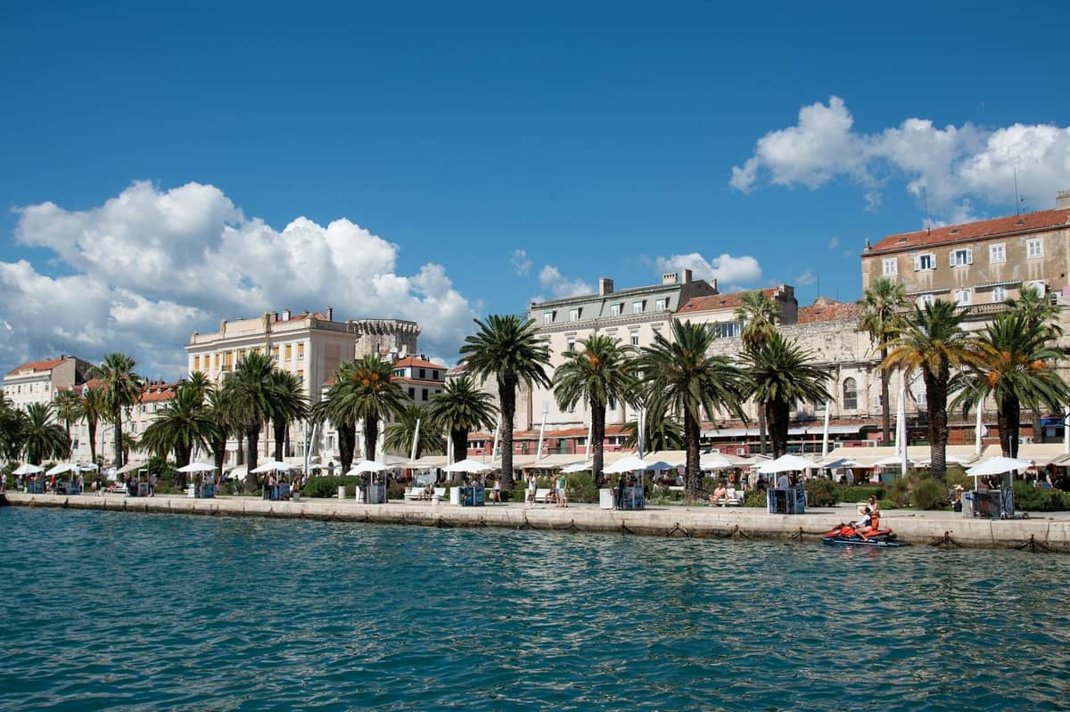 Sonnige Uferpromenade von Split gesäumt von Palmen, historischen Gebäuden und Straßencafés unter einem blauen Himmel mit vereinzelten Wolken.