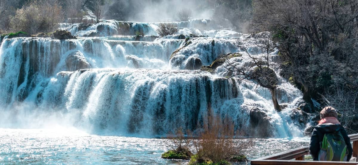 Mehrere Stufen der Wasserfälle des Nationalparks Krka stürzen über felsige Vorsprünge mit aufsteigendem Nebel, umgeben von kahlen Winterbäumen und fließendem Wasser.