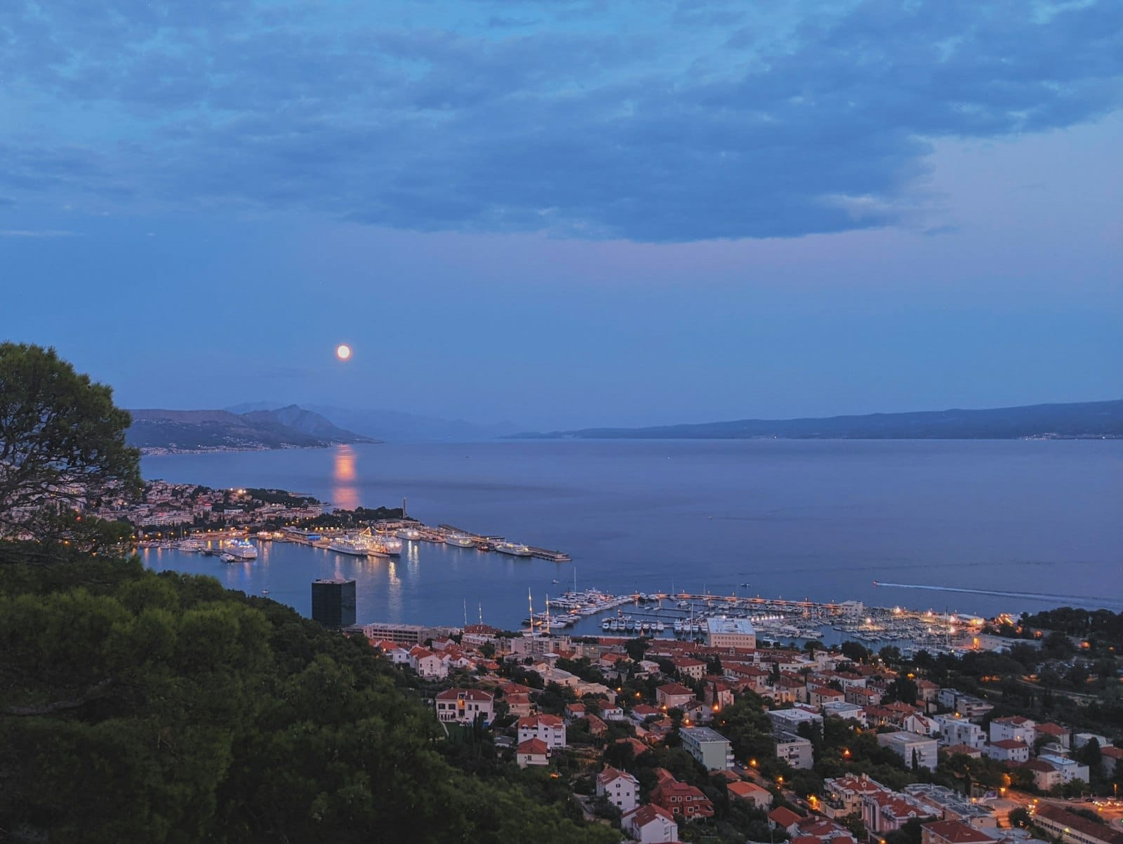 Vista aérea de Split, Croácia, ao entardecer, com as luzes da cidade, o porto e o reflexo da lua no Mar Adriático, transmitindo a energia vibrante da vida noturna.