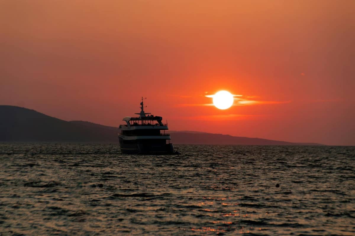Silhouette eines Bootes vor einem dramatischen Sonnenuntergang über dem Meer, mit hügeliger Küstenlinie in der Ferne und orangefarbenem Himmel.