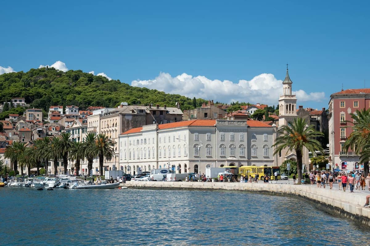 Tagesansicht der Riva-Promenade in Split, Uferpromenade, Palmen und historische Gebäude mit Spaziergängern auf dem Fußweg.