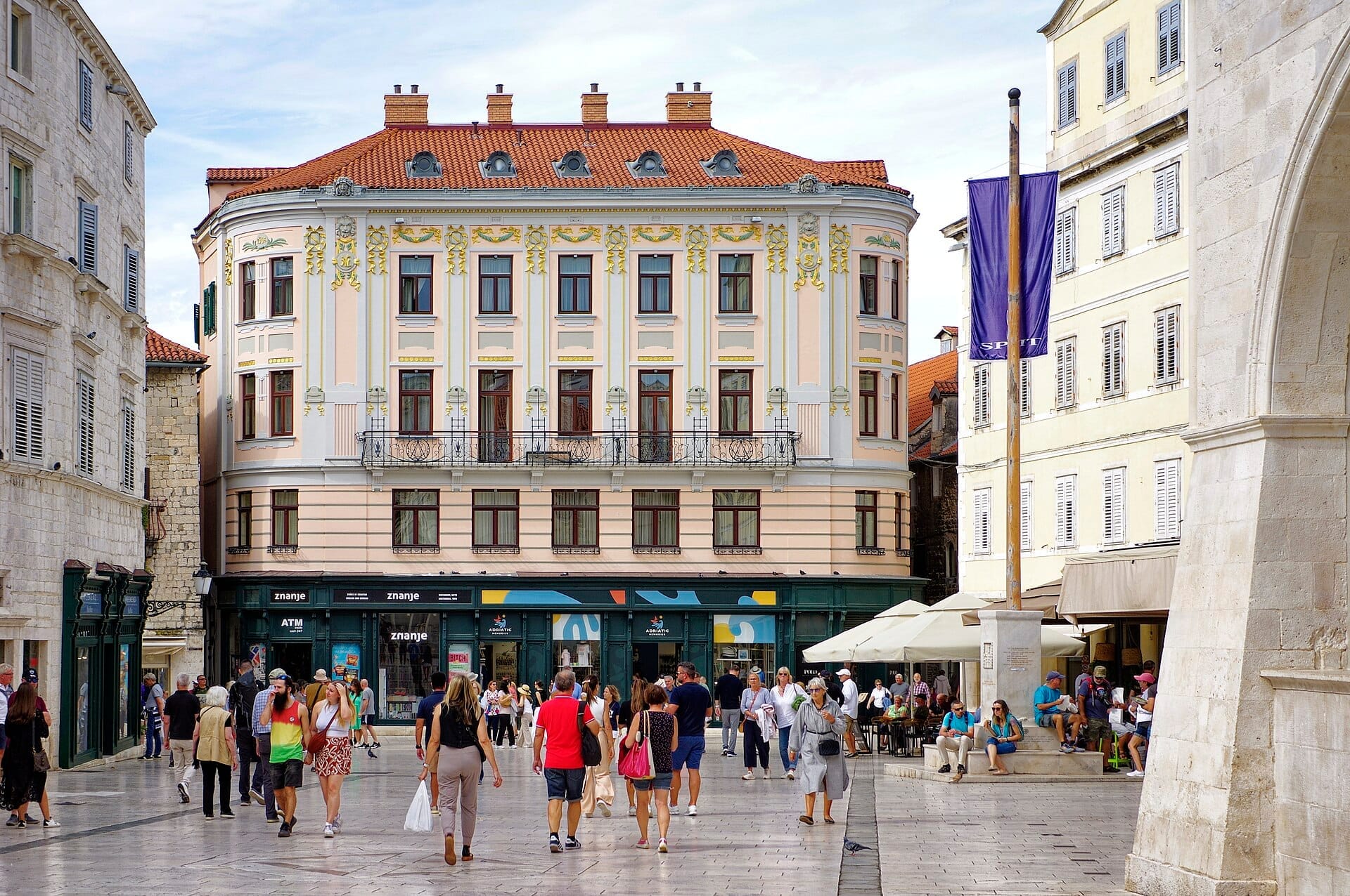Grupos de pessoas caminham e relaxam na histórica praça principal de Split, rodeada de ornamentados edifícios em tons pastel com telhados vermelhos sob um céu luminoso.